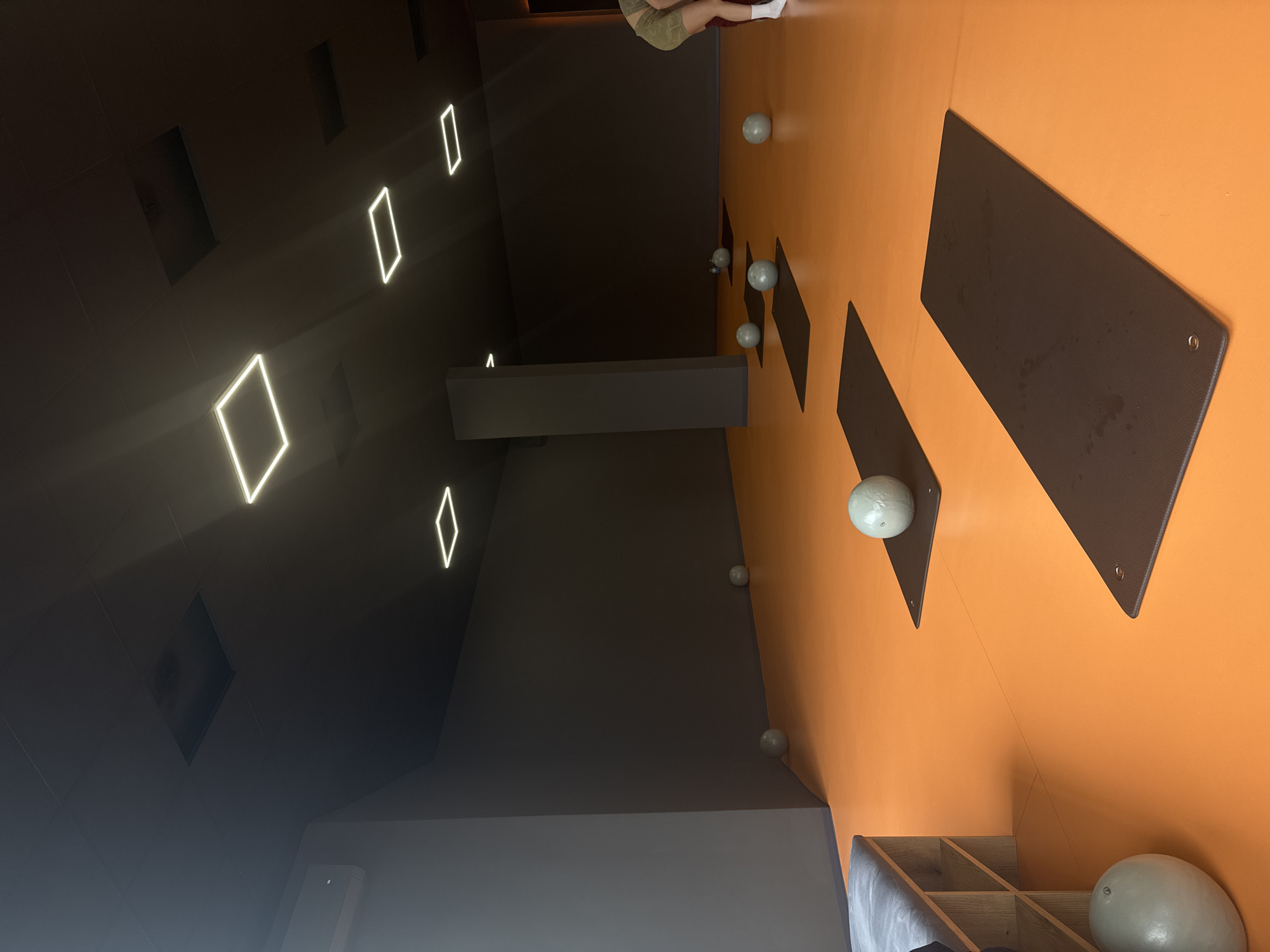 The image shows an interior space in Goce Delcev, Bulgaria. The room is designed with an orange floor and a dark gray ceiling. There are several rectangular and square-shaped light fixtures embedded in the ceiling. A structural element or beam, also dark gray, extends from the ceiling down the middle of the image. On the orange floor, there are several dark-colored rectangular mats and white exercise balls scattered around. A person is partially visible in the top center of the frame. A storage unit with shelves is visible in the lower right corner, containing additional exercise balls.