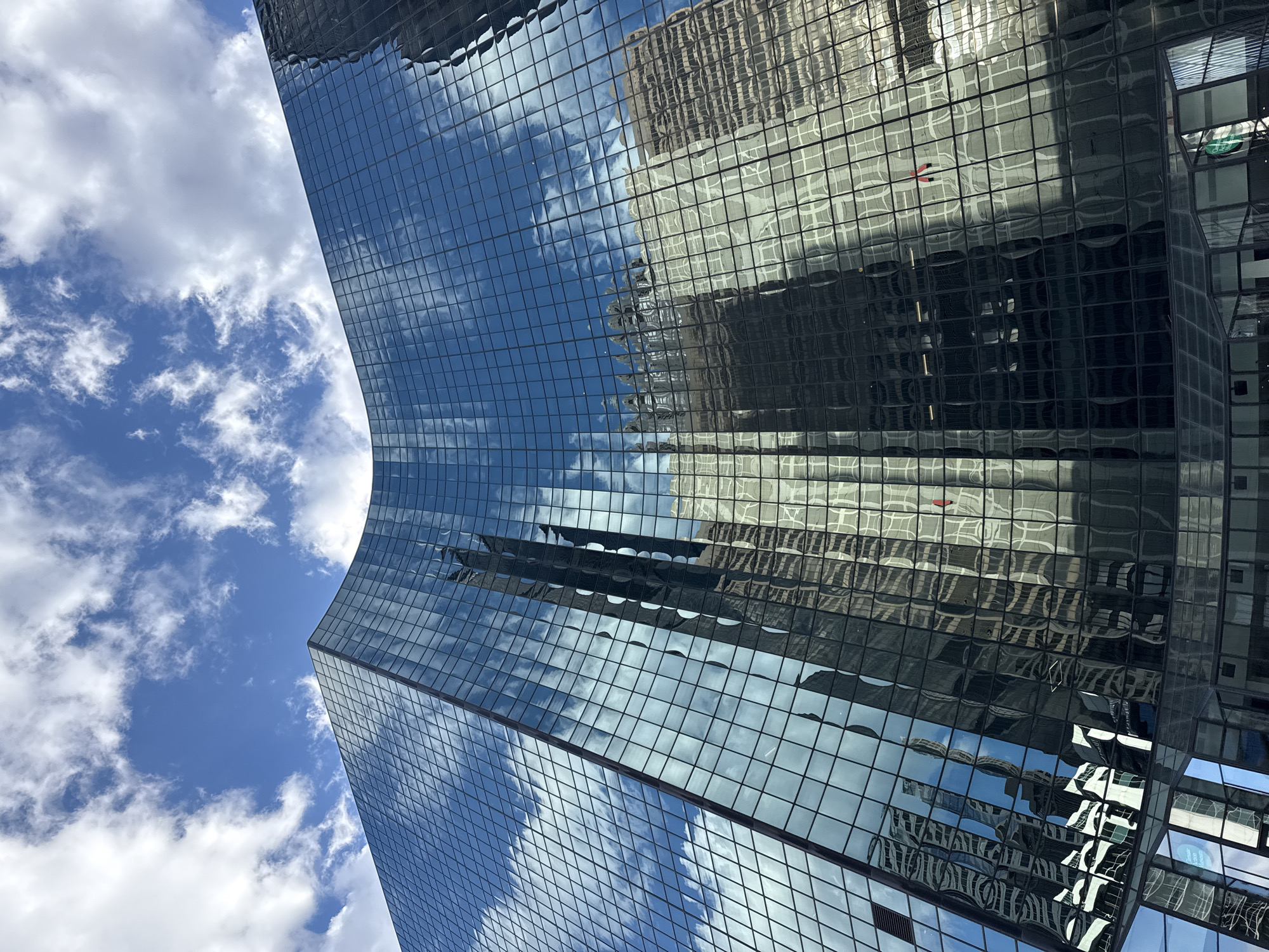 The image shows a portion of a glass skyscraper in Chicago, USA. The building's glass facade reflects the sky, clouds, and other nearby buildings. The sky is blue with white clouds. The architectural style is modern, characterized by clean lines and geometric shapes. The photograph is taken from a low angle, emphasizing the height and scale of the skyscraper.