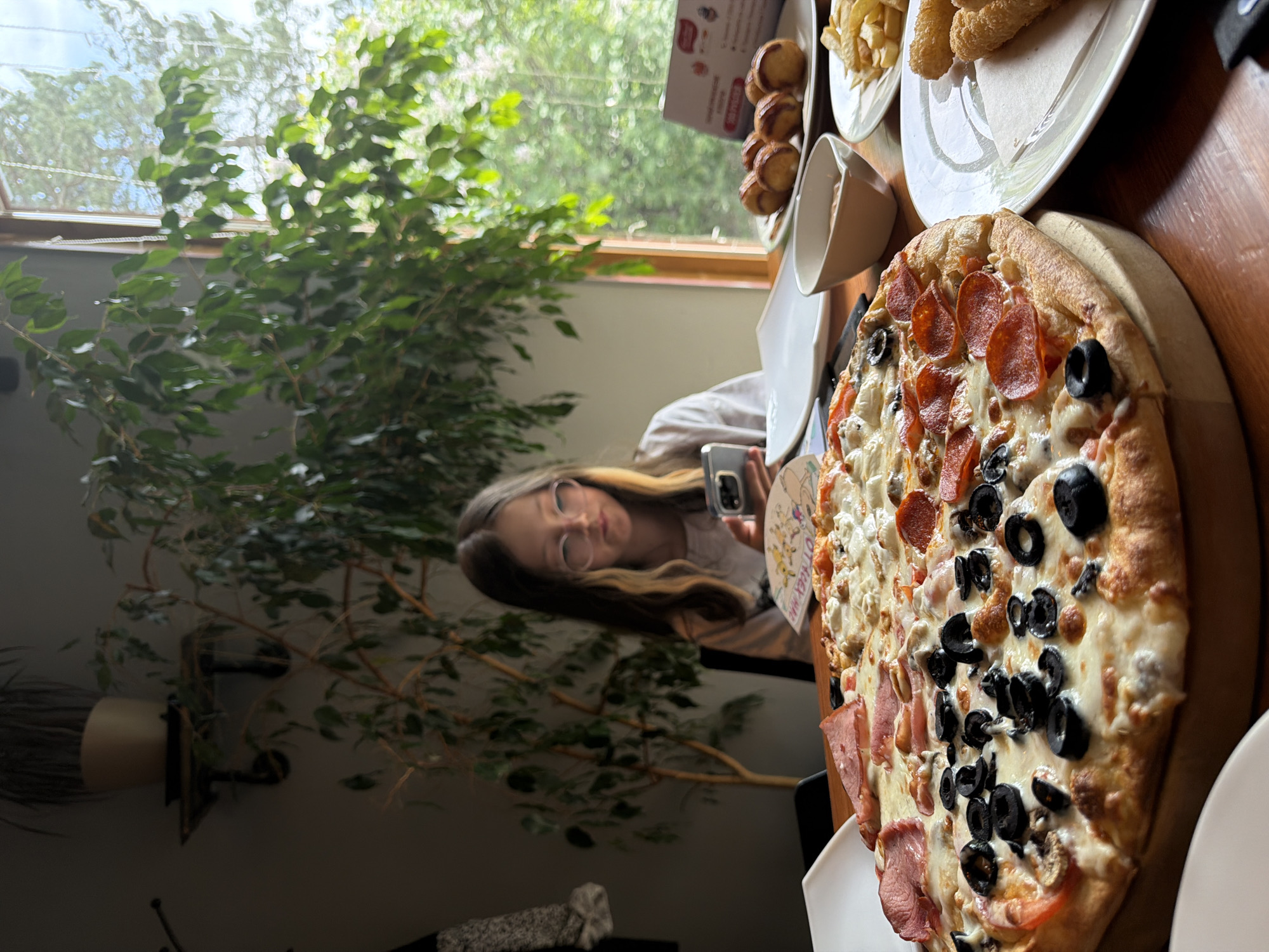 A person is seen in a seated position at a table in an indoor environment, possibly a restaurant or home, in Birobidzhan, Russia. The table is laden with food, including a pizza topped with pepperoni and black olives. Other food items are visible, such as fries and what appears to be fried dough. The person is holding a smartphone and may be taking a picture of the food. A tree-like plant is located near the window.