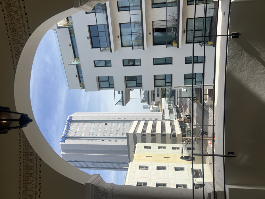 The image depicts an urban street scene in Tel Aviv-Yafo, Israel, viewed from an arched balcony or window.
In the immediate foreground, a light-colored arched architectural element frames the upper portion of the scene. A dark, ornate lantern hangs from the center of this arch, and decorative patterns are visible on its underside. A black metal railing crosses the lower third of the image, indicating a balcony viewpoint, with a light-colored structural pillar partially visible on the left. Shadows are cast by the railing onto the balcony's wall.
The middle ground shows a city street with pedestrian walkways and vehicular markings, including a red and white striped curb. On the left side of the street, two individuals are standing near a utility pole, and further down, another individual on a scooter or motorcycle is visible, moving left to right. Graffiti is present on a lower wall section to the left. On the right side of the street, a white security camera is mounted on a pole.
The background consists of several buildings under a clear blue sky with minimal scattered clouds.
To the left, a tall, light grey, multi-story building with "Metropolitan" written in blue letters near its top identifies the Metropolitan Hotel Tel Aviv. Below it, a beige/yellowish building with multiple windows, followed by a building with horizontal window strips, line the street.
To the right, a modern, white, multi-story residential or commercial building features numerous balconies with glass railings and dark frames. Some balconies contain outdoor furniture, such as chairs and tables, and yellow potted objects. A yellow ladder is visible on a balcony on the third level from the bottom. The rooftop of this building has visible green plants and a glass barrier. Distant buildings are visible on the horizon.