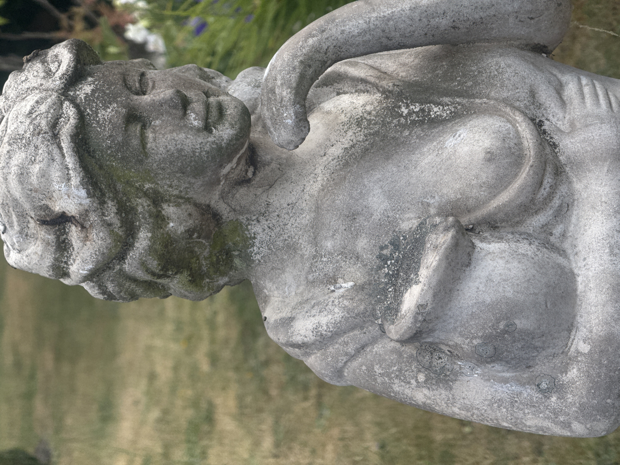 A weathered, grey stone statue of a woman is positioned outdoors, against a blurred backdrop of green grass and foliage. The statue depicts the woman from the chest up, with her head slightly tilted to her right and her right arm bent, with her hand resting on her chin. Her hair is styled in an ornate updo. The stone shows signs of age, with patches of moss and discoloration, suggesting it has been exposed to the elements for a significant period. The lighting suggests it is daytime. The statue is located in Heidelbach, Germany.