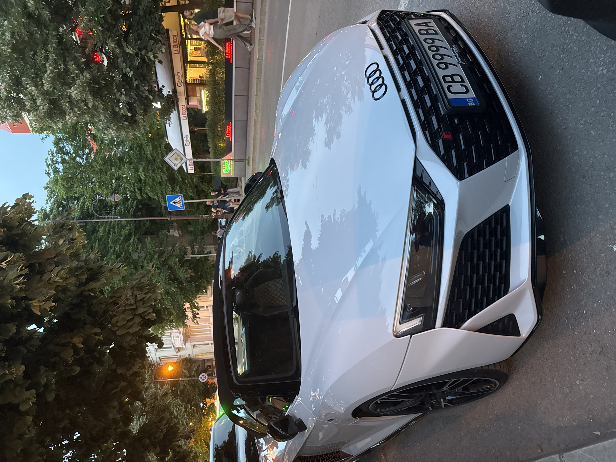 A white Audi R8 convertible is parked on a street, with its top down. The car is the main subject, positioned in the foreground and angled slightly towards the right. The vehicle features a black grille, black rims, and a black soft top. The license plate is visible and reads "CB 9999 BA". In the background, there are trees, a building with several shops and restaurants, and pedestrians walking on the sidewalk. The sky is overcast, suggesting it might be late afternoon or early evening.