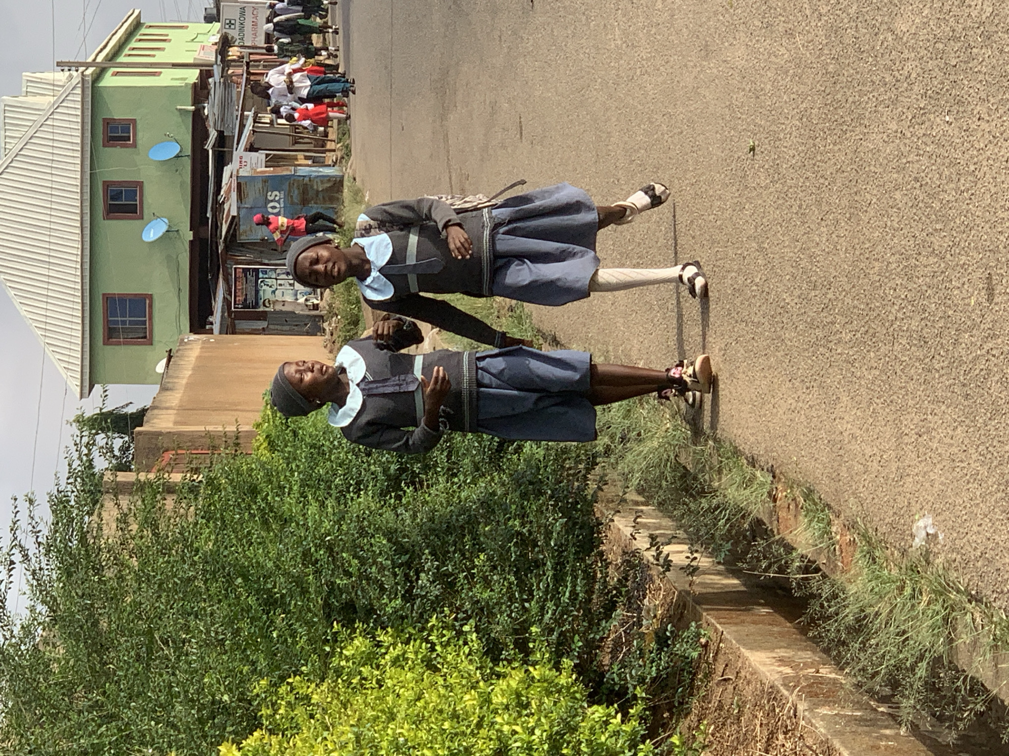 Two schoolgirls in grey and blue uniforms stand on a street in Jos, Nigeria. They both wear grey head coverings and sandals. One girl has a single white sock, and the other has on black hosiery. The street is paved and runs alongside a grassy shoulder and a concrete drainage ditch overgrown with greenery. A green building with a white roof and blue satellite dishes is visible in the background, along with people congregating on the other side of the road. A sign reading "DADINKOWA PHARMACY" is also visible.