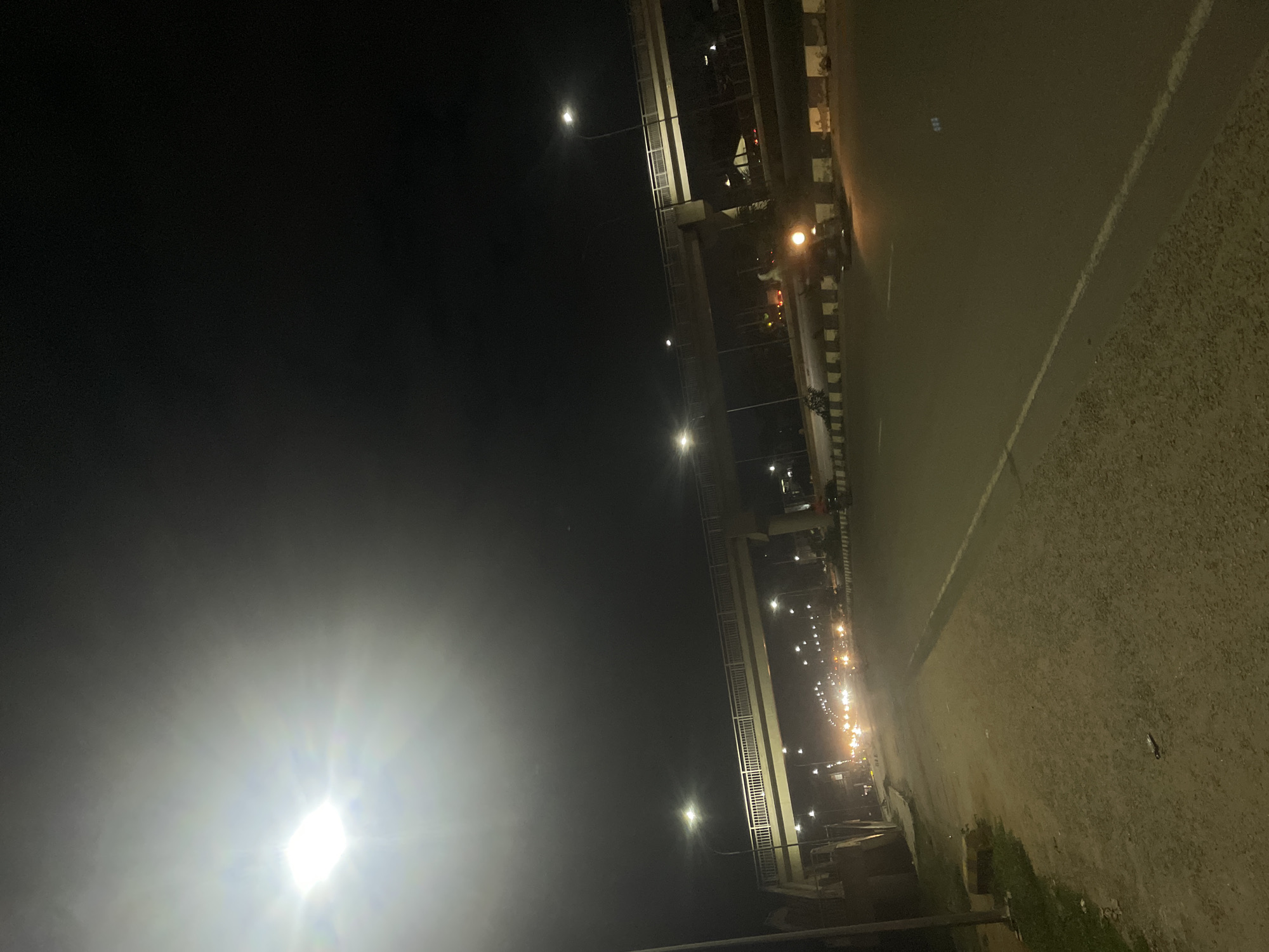 The image shows a street scene at night in Abuja, Nigeria. A pedestrian bridge spans across a multi-lane road, illuminated by streetlights. Traffic, consisting of cars and possibly motorcycles, is visible on the road, their headlights and taillights creating streaks of light. The road surface is asphalt, with white lines marking the lanes and a rougher, unpaved shoulder on the left side of the image. Along the roadside, there are decorative checkered barriers. The sky is dark, with a bright, overexposed streetlight in the upper left quadrant of the image. The overall impression is of an urban environment at night, with infrastructure for transportation and lighting.