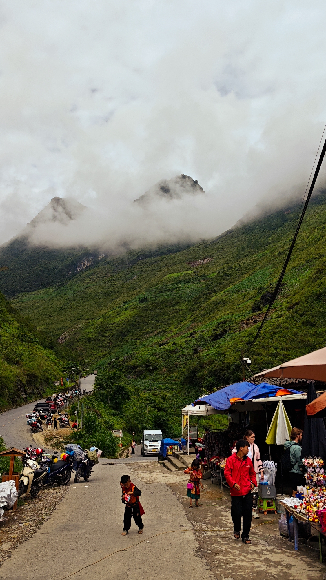 The image depicts a street scene in Yên Minh Commune, Vietnam. The environment is characterized by lush green mountains partially shrouded in clouds. The street is lined with parked motorbikes and a row of market stalls under blue and brown awnings. Several people are present: a young child in traditional clothing is walking on the street, another young child is standing near the market stalls, and adults are browsing at the stalls. A white van is parked further down the street. The overall atmosphere suggests a rural or mountainous area with a local market.