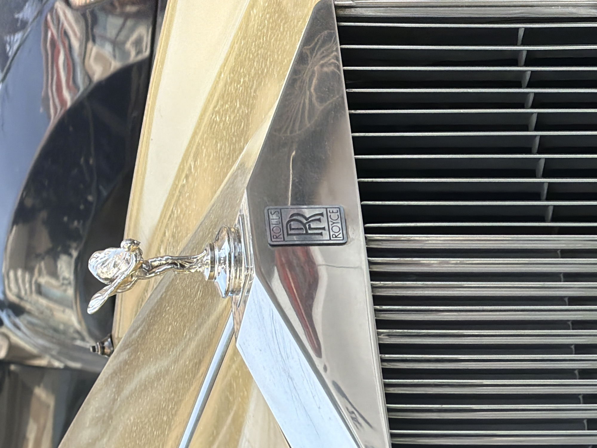 The image displays a detailed, close-up, frontal view of the hood and upper grille of a luxury automobile, specifically a Rolls-Royce. Dominating the center is the silver-toned "Spirit of Ecstasy" hood ornament, positioned atop a polished chrome section. Below the ornament, a rectangular Rolls-Royce badge with the iconic double 'R' emblem is centrally affixed to the chrome radiator surround. A vertical-slatted chrome grille extends downwards from this section. The vehicle's hood is finished in a light gold or champagne metallic paint, exhibiting a visible layer of dust and dirt, with distinct streaky patterns. In the upper right background, a darker, possibly navy blue, section of the vehicle is visible, showing reflections, including a distinct pattern of red, white, and blue stripes consistent with the Bulgarian national flag. No individuals or active interactions are present. The scene is static, captured in daylight conditions, and is situated within the city of Varna, Bulgaria.