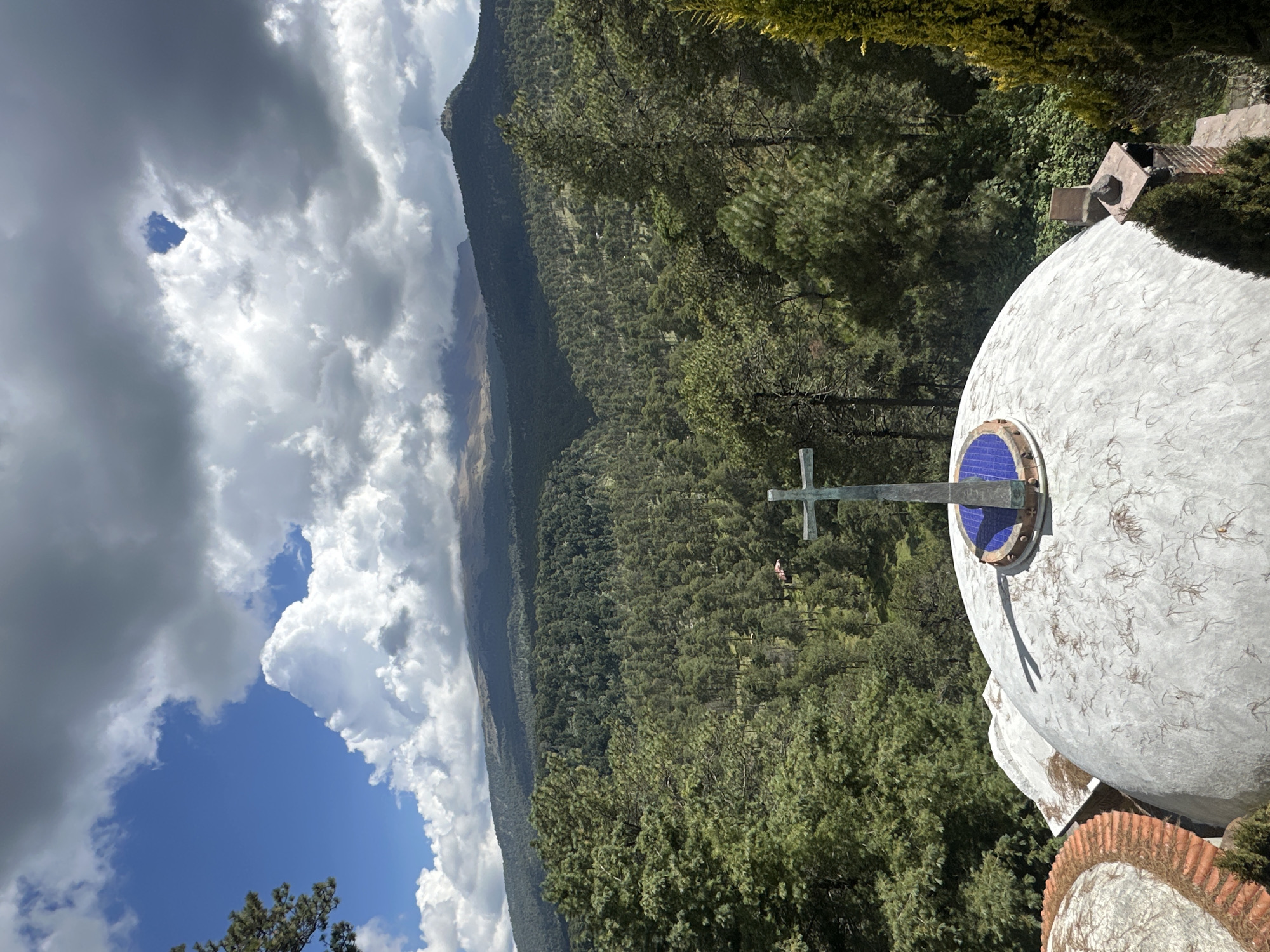 The image captures a scenic view from a building with a domed roof, featuring a prominent cross atop the dome. The dome is white and appears weathered, with some dried leaves scattered on its surface. Adjacent to it, another part of the structure has a terracotta tiled roof.
The background is dominated by a dense forest of pine trees that covers rolling hills. In the far distance, a mountain range is visible, partially obscured by clouds. The sky is a mixture of blue patches and dramatic, heavy clouds, suggesting a dynamic weather system. A small, red-roofed building is nestled among the trees in the middle ground.
The perspective suggests the viewpoint is elevated, looking out over the landscape. The lighting indicates it is daytime, with shadows cast on the dome from the cross and other unseen objects. The overall scene conveys a sense of natural beauty and tranquility, possibly from a religious or historical site in the region of San Mateo Ozolco, Mexico. No people are visible in the image.