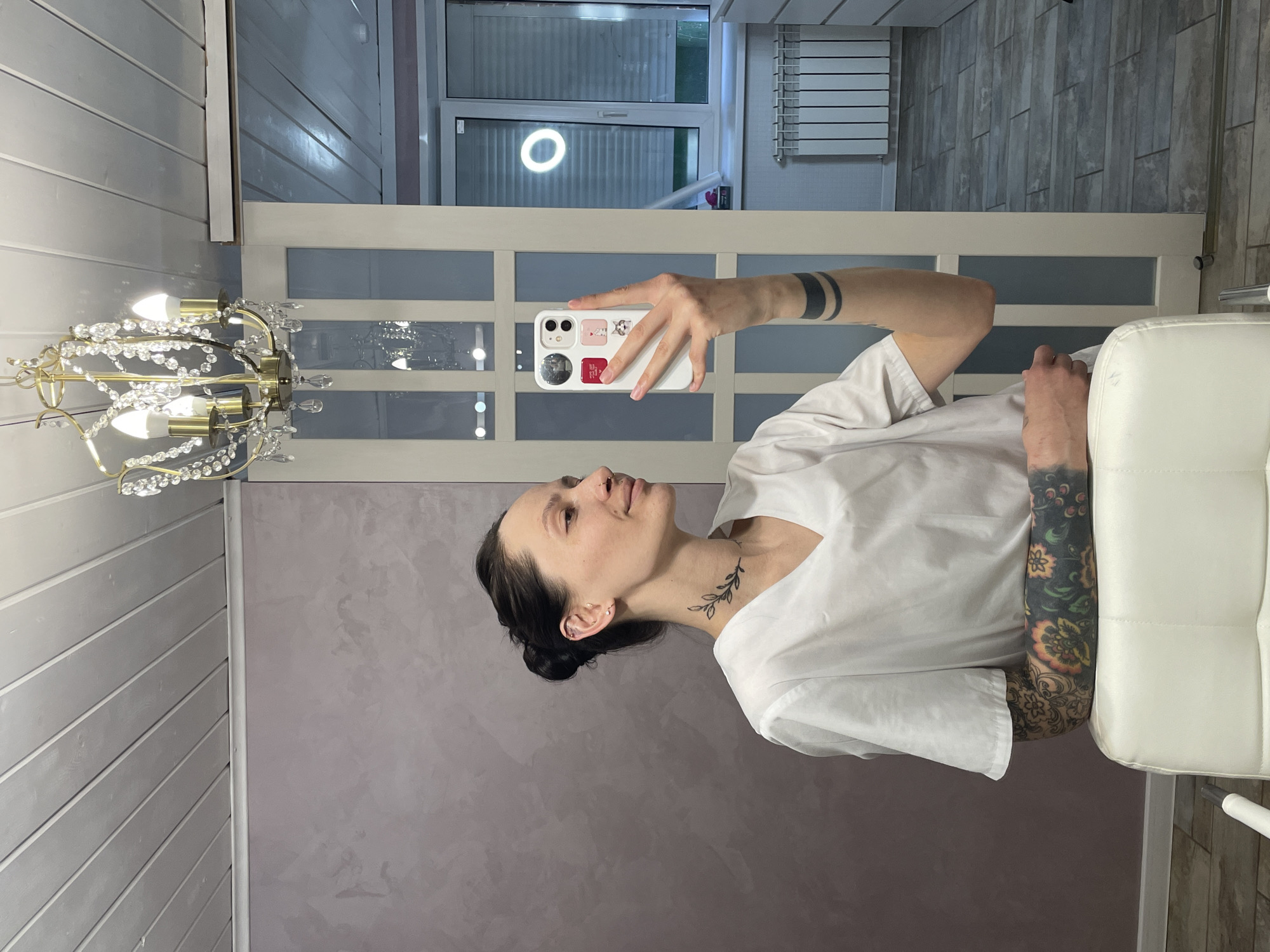 A woman with dark hair pulled back in a bun takes a selfie in what appears to be an indoor setting. She is wearing a white t-shirt. Her visible arm is adorned with tattoos. She holds a white smartphone in her hand. The background includes a chandelier, a door with frosted glass panes, and part of a window. The wall behind her is a muted purple or lavender color. There are no visible indications that place the location in the city of Izhevsk, Russia.
