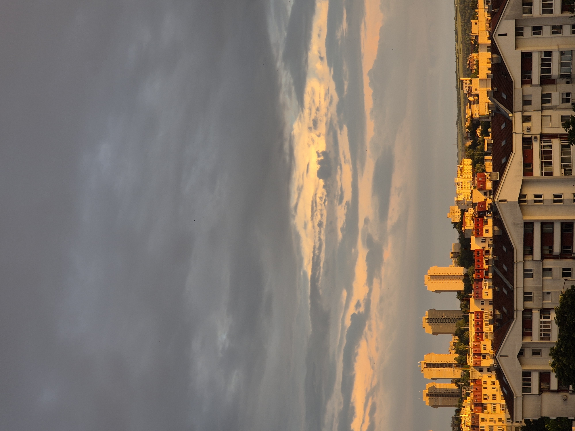 This vertical image depicts an urban landscape within the city of Unknown, Unknown, captured during what appears to be either sunrise or sunset. The upper two-thirds of the frame are dominated by an overcast sky, with dense, dark grey clouds at the top transitioning to lighter grey and warm orange-yellow hues near the horizon where sunlight penetrates the cloud cover. Numerous small, dark specks, identified as birds, are scattered across the illuminated and darker sky sections.
The lower third of the image showcases a dense arrangement of buildings. In the mid-ground, four prominent, tall, light-colored (likely concrete) multi-story buildings, characterized by their blocky, angular designs, stand silhouetted and illuminated by the warm, golden light. These are surrounded by numerous smaller, multi-story residential buildings, also bathed in the same golden light, featuring red or orange-accented balconies and roof elements. The immediate foreground reveals the upper sections and roofs of additional residential buildings. These structures are predominantly white or light-toned, with dark, sloped tiled roofs and numerous windows and balconies. There are no discernible people or explicit actions captured within the scene; the focus is on the urban environment under specific lighting conditions.