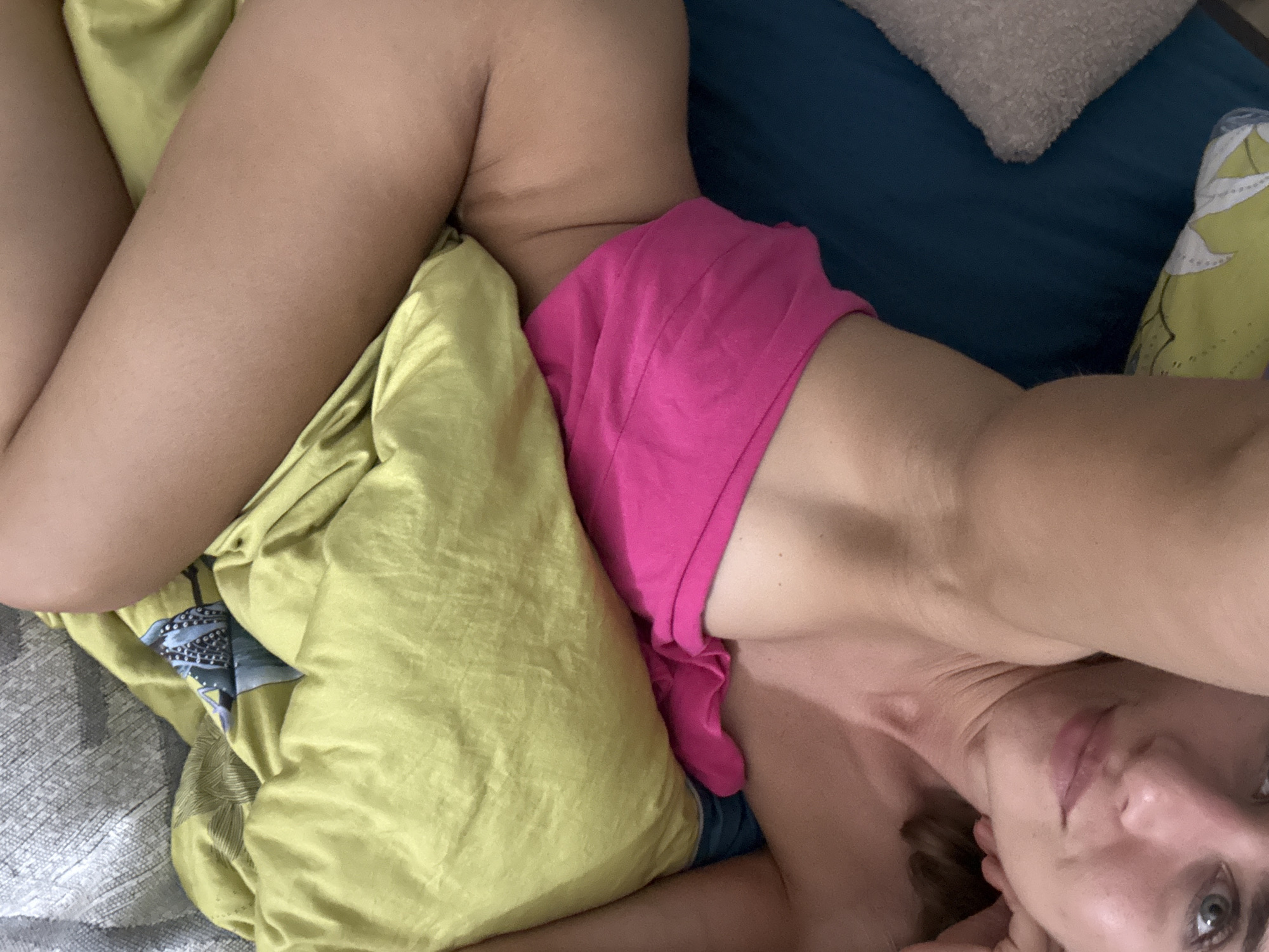 The image shows a woman lying on a bed. She is wearing pink shorts. The bed is covered with a yellow sheet and a blue pillow. There is also a gray pillow and a pillow with a floral pattern visible. The woman is looking up at the camera and smiling. The lighting in the image suggests that it is daytime. The overall mood of the image is relaxed and intimate.