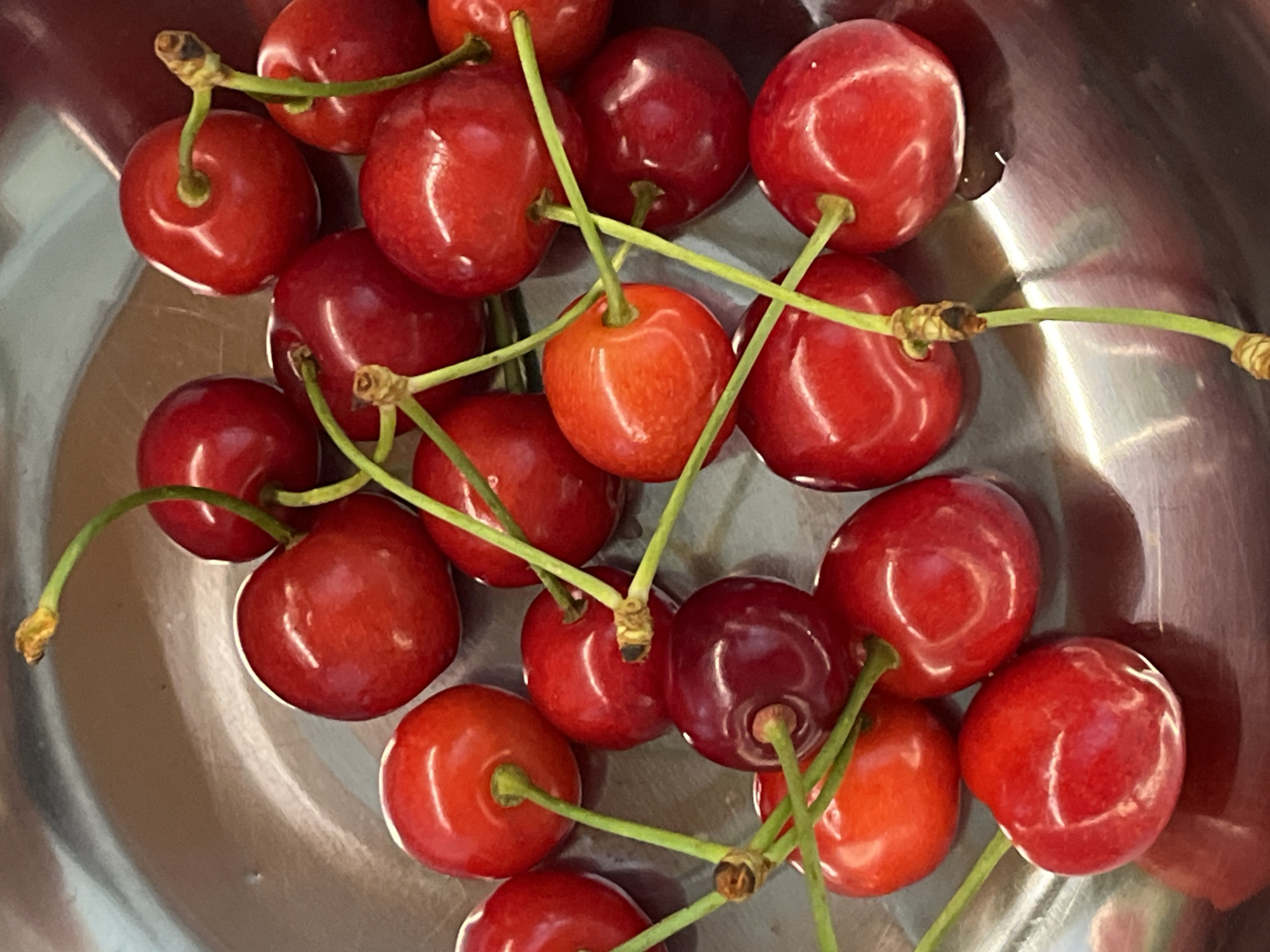 This is a close-up, overhead shot of a pile of fresh, red cherries in a metal bowl. The cherries are plump and glossy, with visible stems attached to some. The lighting creates highlights on the surface of the fruit, suggesting it might be indoors or in an area with artificial light. The arrangement of the cherries is natural and somewhat random, with some overlapping. The metallic sheen of the bowl is visible around the edges of the frame, indicating a reflective surface. No people, text, or specific location cues are apparent in the image. The focus is entirely on the texture, color, and form of the cherries.