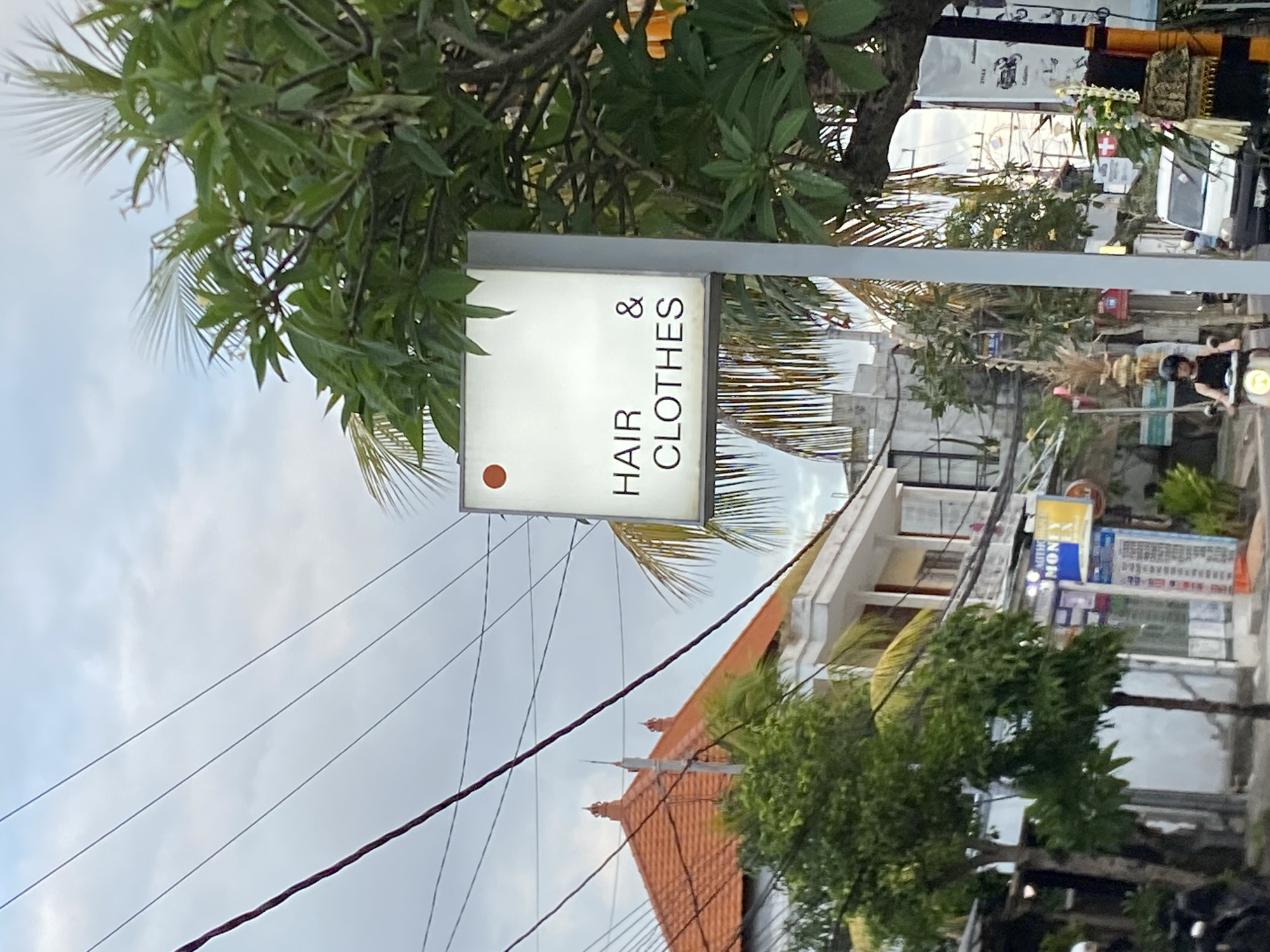 In Mengwi, Indonesia, an outdoor scene captures a street view with a prominent illuminated sign displaying "HAIR & CLOTHES" with a small orange dot above the text. The sign is mounted on a white pole and partially obscured by lush green foliage from a nearby tree. To the left, a traditional Indonesian building with an orange tiled roof and white walls is visible, with electrical wires crisscrossing the sky. Several other buildings and shops line the street, with some signage indicating businesses such as "MONEY EXCHANGE". A few motorcycles are parked, and one person is riding a motorcycle in the foreground. The sky is overcast, suggesting a cloudy day. The overall environment depicts a commercial street in a tropical locale.