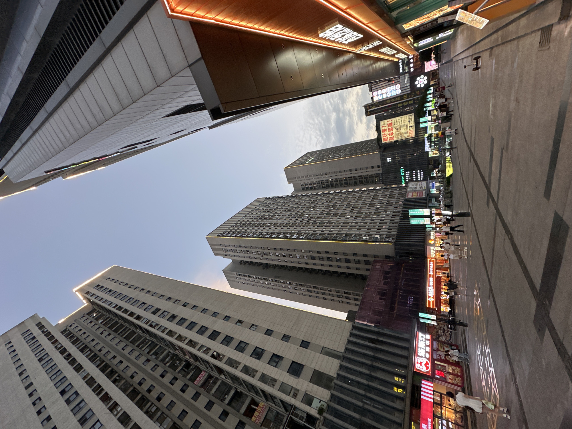 The image, taken from a low angle, depicts a street scene in the Chongqing High-tech Zone, China. Several high-rise buildings frame the sky. On the right side of the image, storefronts are visible, illuminated by bright signage. Pedestrians walk along the sidewalk. The street is paved with gray rectangular tiles with dark lines. Signage and advertisements are visible on the buildings. The sky is light blue with a few clouds. Some text is visible in Chinese characters in the store signs. A few people are visible in the street.
