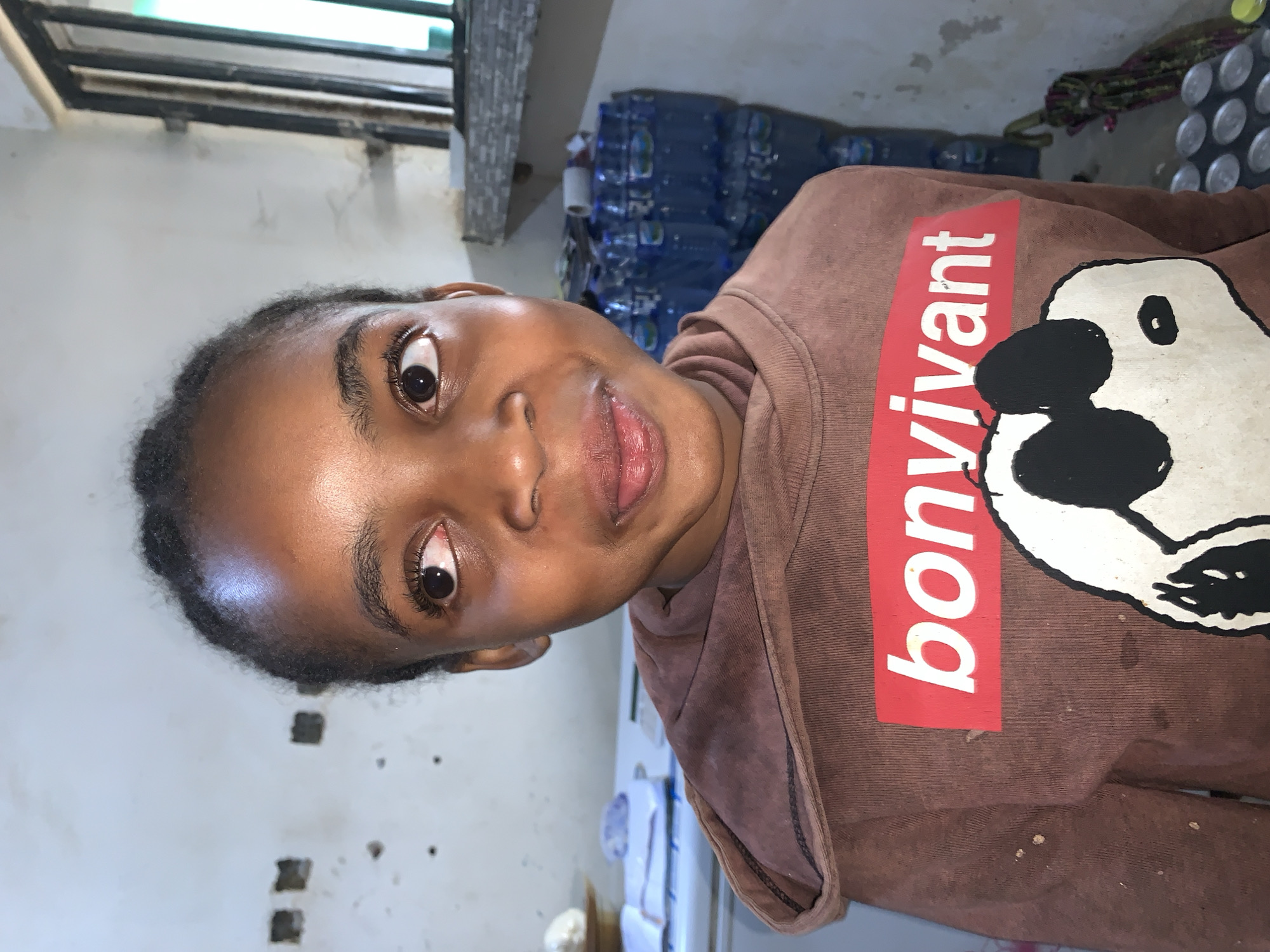 In New Stadium, Nigeria, a young individual with dark skin and braided hair is depicted indoors, looking upwards and to the right with a slightly protruding tongue. The person wears a brown hooded sweatshirt featuring a red rectangular graphic with "bonvivant" in white lettering, positioned above a large black and white cartoon-style character's head. Several light-colored stains are visible on the sweatshirt. The background to the left reveals a light-colored wall with multiple dark marks and two square-shaped indentations, above a white surface displaying a light object, a dark object, and a white paper booklet. To the right, a window with dark vertical bars shows a hint of green beyond, situated above a grey tiled surface. Below this, a substantial stack of clear plastic water bottles with blue caps is visible, resting on a floor that also shows several empty silver cans and a colorful, twisted fabric object.