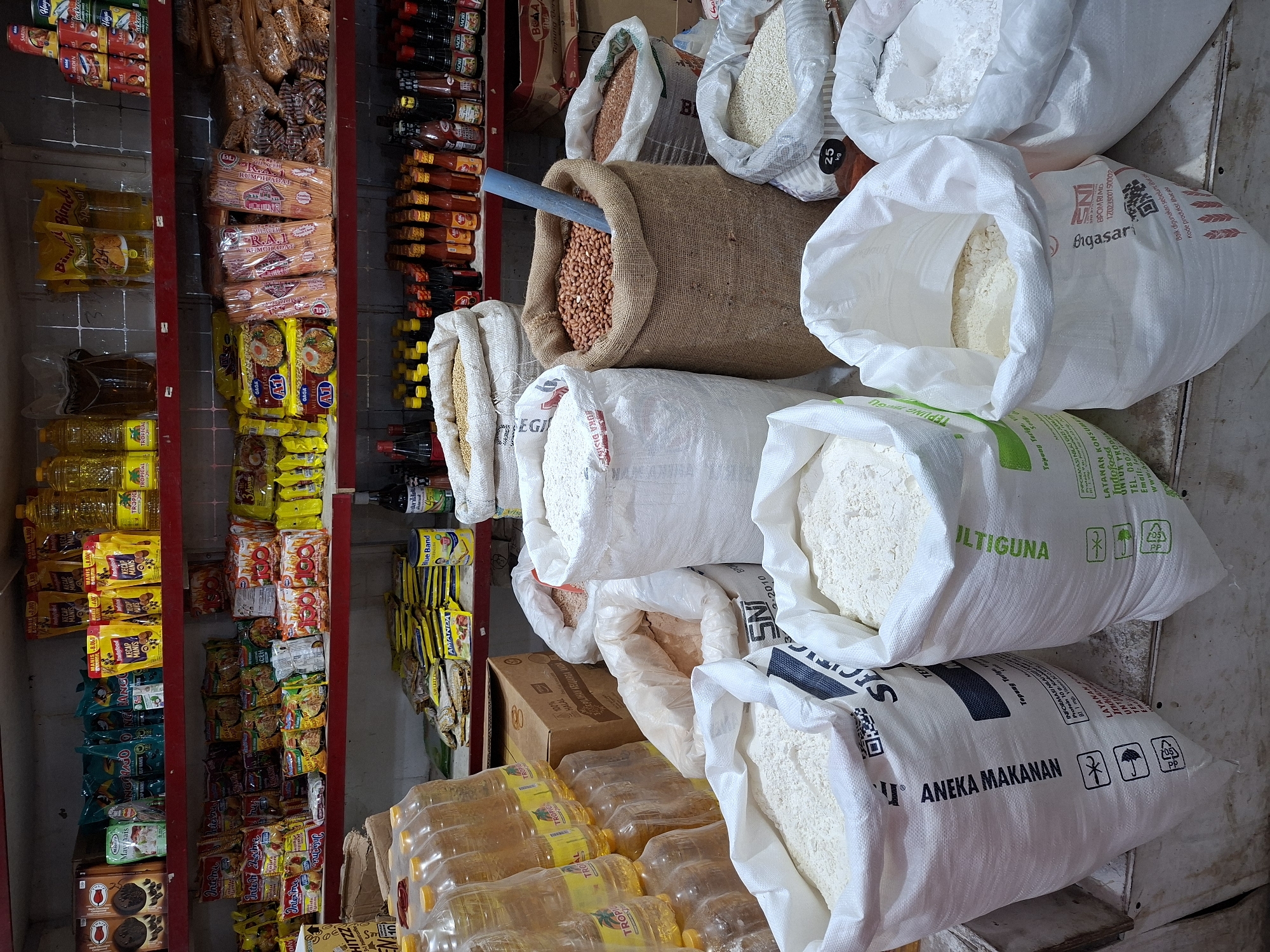 This image shows the interior of a small grocery store in Lhokseumawe, Indonesia. A red metal shelving unit on the left holds various packaged goods, including instant noodles (Indomie and Gaga), sauces, cooking oil, and snacks. In the center and to the right are large bags of flour, rice, and peanuts. The bags are of various brands, including "Bigasari," "Tepung Terigu," and "Aneka Makanan." The store appears to be well-stocked with essential food items. The lighting is somewhat dim, and the shelves are tightly packed with merchandise.