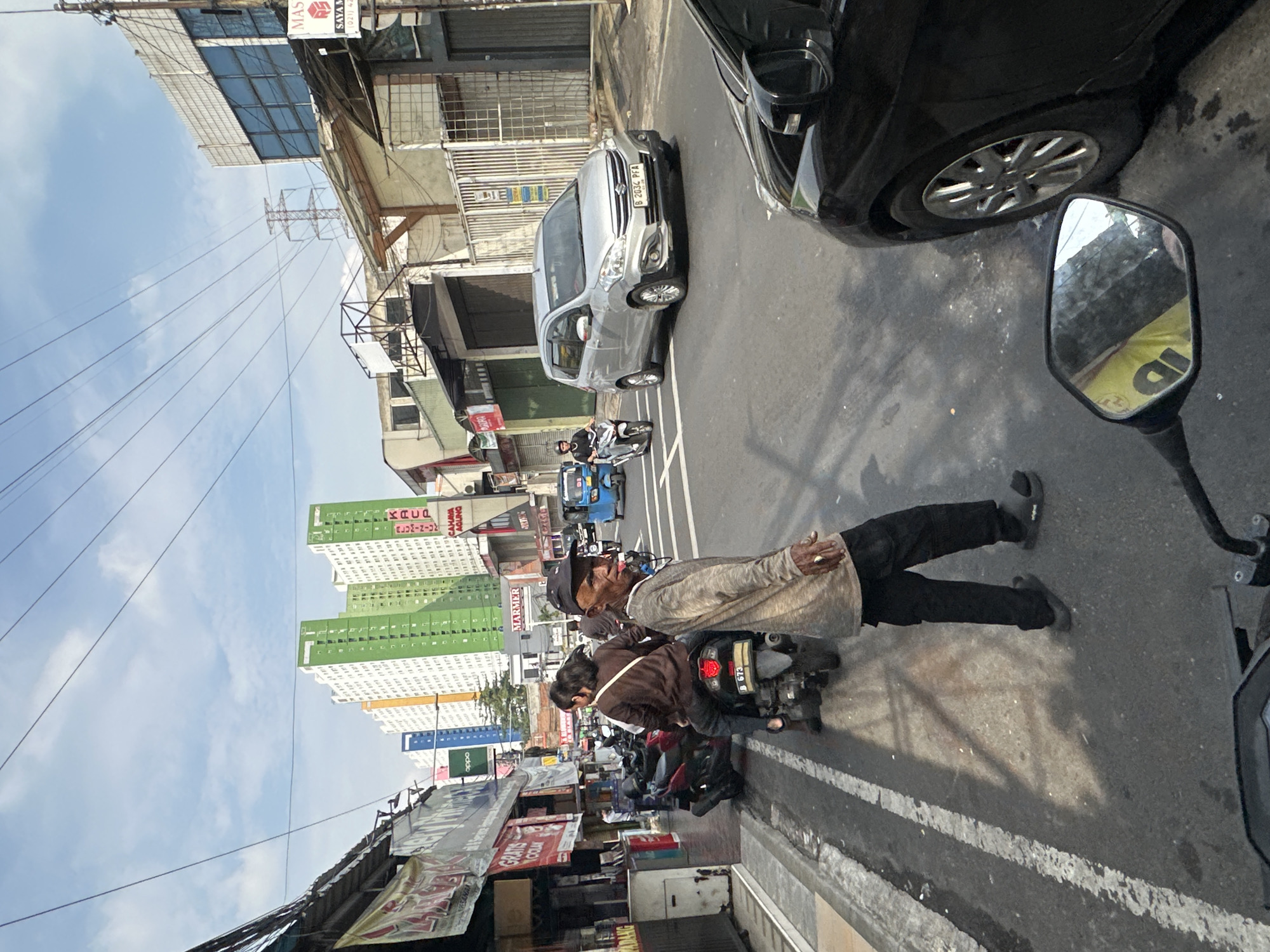 A medium shot depicts a street scene in Central Jakarta, Indonesia. The image is taken from the perspective of someone on a motorcycle, with the motorcycle's mirror visible in the foreground. An older man is seen riding on a motorcycle, with another person behind him. A line of parked motorcycles are visible along the side of the road. Several buildings are in the background, including a tall green and white apartment complex. Cars are also visible on the street, including a silver Suzuki Ertiga with license plate B 2034 PFA. Various signs and advertisements are visible on the buildings, including ones for Oppo and Kacap. The sky is partly cloudy.