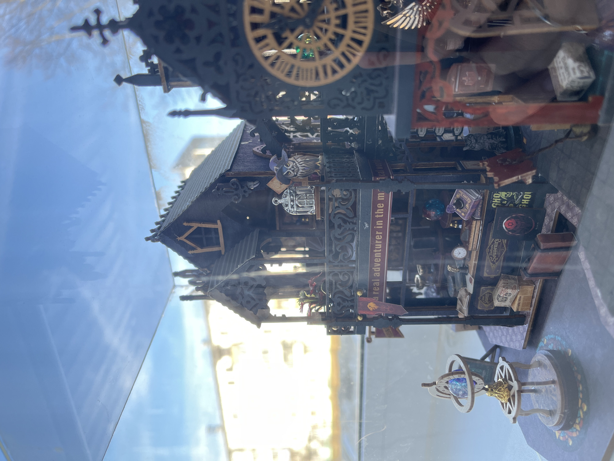 The image displays a detailed miniature diorama, enclosed within a glass case, situated in the city of Chaikovsky, Russia. The central subject is a multi-story structure rendered in a dark, gothic architectural style, featuring an elaborate pointed roof, a small dormer window, and balconies adorned with intricate railings.
The ground level of the model depicts a storefront with various miniature items, including books (some labeled "GHO"), packages, a small clock, and two brown suitcases. A banner with a lion emblem hangs near a partially visible sign that reads "a real adventurer in the m." A miniature grey cat figurine is positioned within the shop. The first-floor balcony contains a small red plant, a silver birdcage, and an owl figurine wearing a wizard hat. Adjacent to the main building on the left, a small table holds an armillary sphere-like object encasing a blue, crystalline sphere. To the right, a portion of another dark, ornate structure is visible, featuring a large circular element resembling a golden clock face.
The glass enclosure of the diorama reflects a clear blue sky with some clouds and the blurred outlines of sunlit, light-colored buildings in the background, indicating an urban setting under bright daylight conditions.