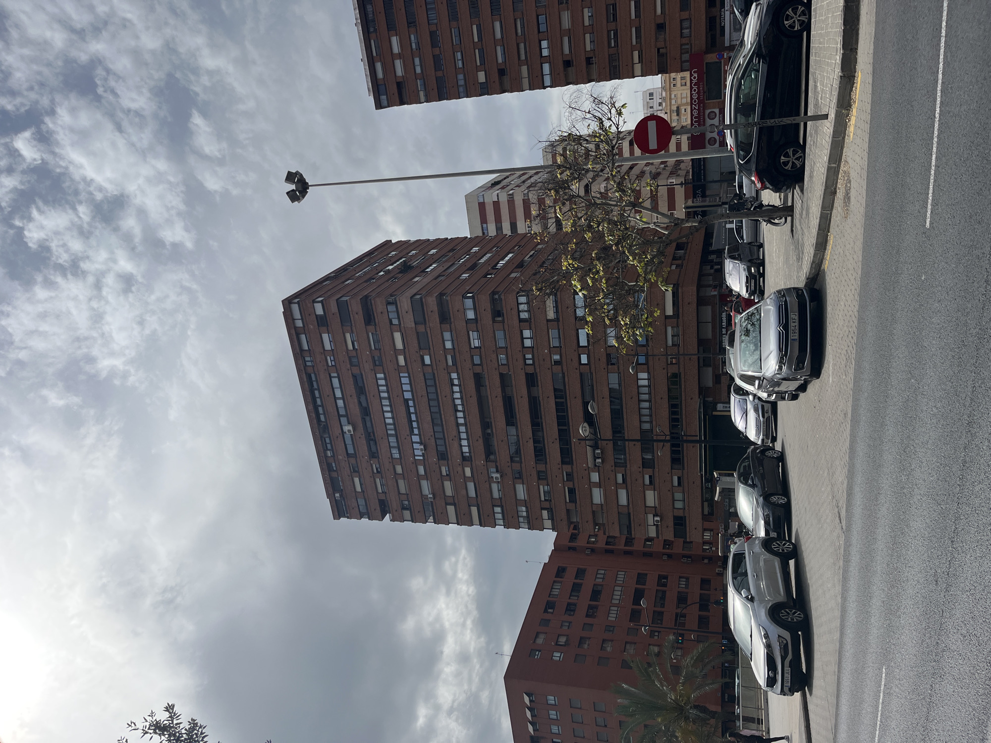 The image depicts a cloudy daytime urban scene in Valencia, Spain, looking up towards tall residential buildings from a street level. Several reddish-brown apartment buildings dominate the background and mid-ground, characterized by numerous windows and some visible air conditioning units. One building further back appears to have horizontal white stripes.
In the foreground and mid-ground, a row of cars is parked along a concrete sidewalk with yellow curb markings, adjacent to an asphalt road featuring a white dashed lane divider. Visible license plates include "1954 KFJ" on a silver car in the immediate foreground and "1651 DLL" on another car further down the street. A black SUV is also parked.
Street furniture includes a tall lamppost with multiple light fixtures and another pole displaying a red circular "No Entry" sign with a white horizontal bar. A deciduous tree with sparse or no leaves stands in the mid-ground, while a palm tree is visible towards the bottom right, suggesting a warm climate.
The sky is overcast with significant cloud cover, indicating a non-sunny day. Visible text on storefronts includes "omeze Cebrian Correduria Seguros" (an insurance brokerage), "NOTARIA", and "Era". No people or explicit activities are visible, but the scene implies a typical urban street environment with residential and commercial elements.