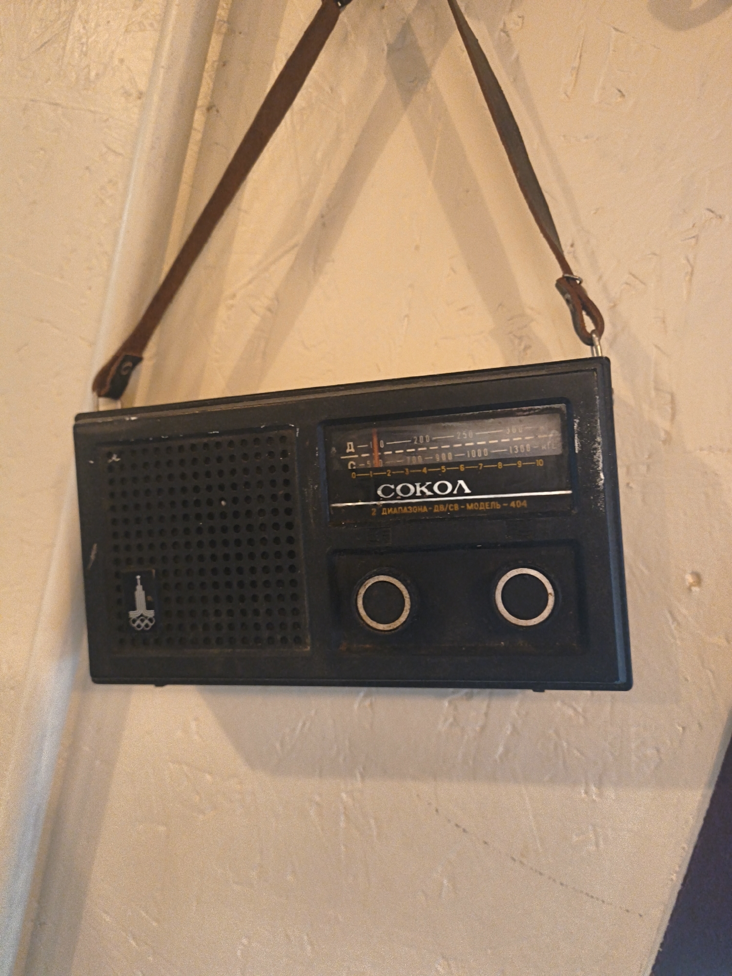 A black, portable transistor radio, branded "SOKOL" with the model number "404" clearly visible on its front, hangs from a brown leather strap against a textured, off-white wall. The radio features a speaker grille on the left, a tuning dial with frequency markings, and two circular buttons or knobs towards the bottom. A small emblem, possibly a stylized torch with Olympic rings, is present on the speaker grille. The strap is attached to the top of the radio and runs up to a hook or point on the wall, creating a triangular suspension. The wall appears to be an interior surface, with subtle shadows and variations in texture indicating it is not a flat, uniformly lit surface. The scene is devoid of people or any indication of specific activity. There is no information within the image to suggest a location in Tallinn, Estonia.
