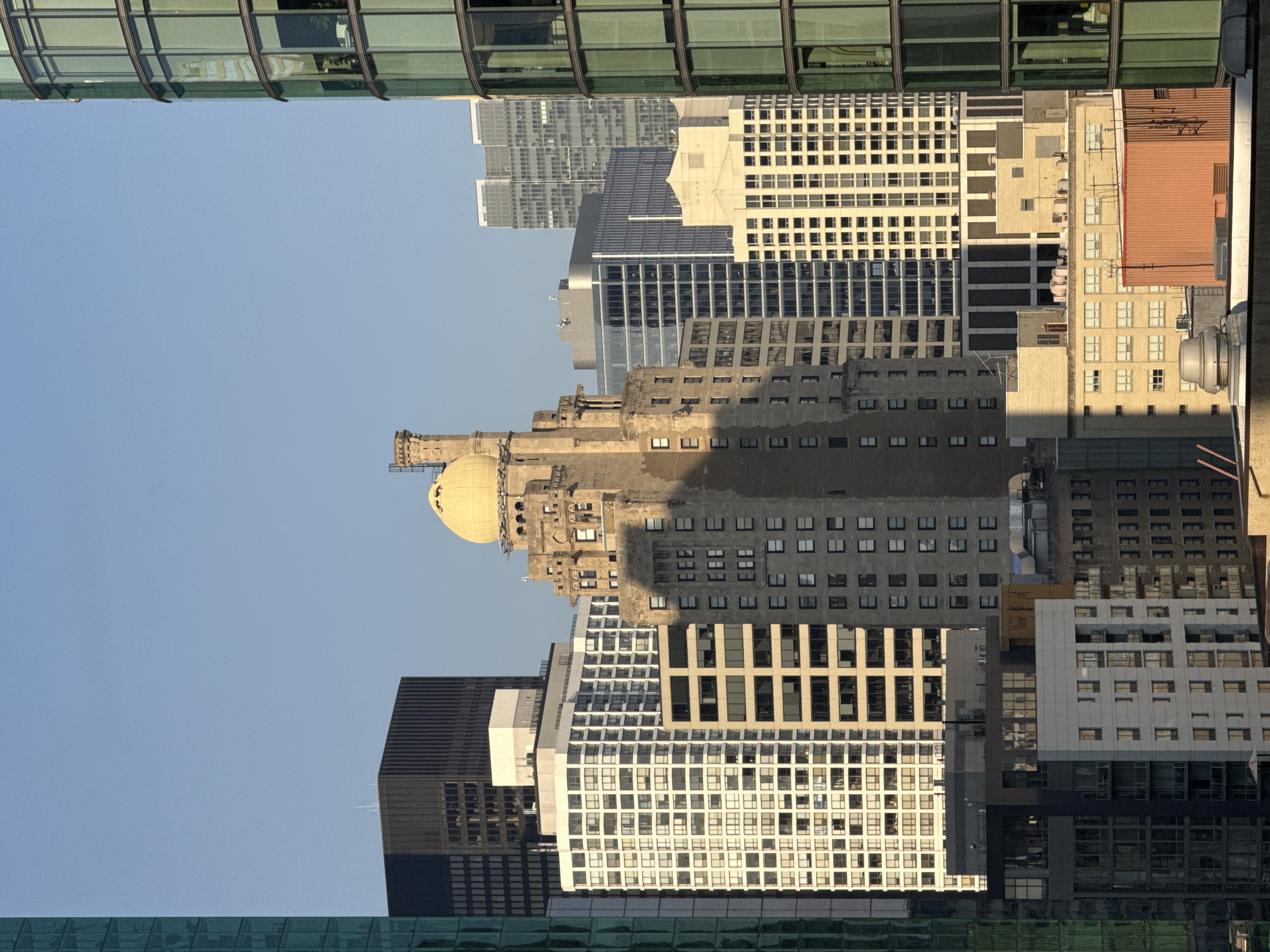 The image depicts a collection of skyscrapers in a city, bathed in the warm light of what appears to be late afternoon or early morning. The sky is a clear, pale blue. The buildings vary in architectural style and material.
Dominating the center of the frame is a distinctive older building with a tan-colored stone facade and ornate detailing. It is topped with a golden dome and a prominent chimney or spire. This building casts a long shadow across its own facade and onto adjacent structures.
To the left of this central building, a tall, white apartment building with numerous windows stands out. Further to the left, partially visible, is a dark, rectangular skyscraper. To the far left, a modern building with a green glass facade is seen.
To the right of the central building, more skyscrapers are visible. One is a light-colored building with a grid of windows. Further to the right, a curved building with a green glass facade, similar to the one on the far left, rises. In the background, several other modern glass and steel buildings can be seen, some appearing taller and more distant.
At the lower part of the image, the roofs of some lower buildings are visible, showing various vents, pipes, and rooftop structures. One building in the foreground has an orange, corrugated metal facade.
The scene is devoid of any visible people or vehicles, suggesting a focus on the urban architecture and the interplay of light and shadow on the city skyline. The overall impression is one of a dense urban environment with a mix of historical and contemporary architectural styles.
While the prompt requests the location to be Chicago, United States of America, there are no specific landmarks or identifying features in the image that definitively confirm this location without external knowledge. However, the style of architecture is consistent with major American cities.