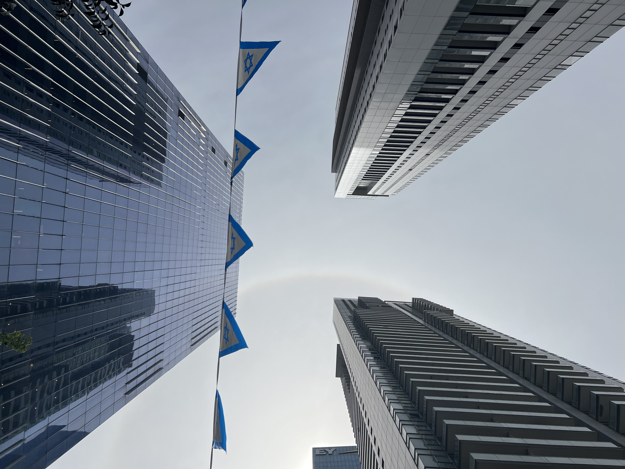 The image is a low-angle shot of three buildings in Tel Aviv, Israel. A string of Israeli flags hangs vertically on the left side of the frame between the two left-most buildings. The building on the far left is a blue glass skyscraper with a grid pattern. To the right of it and slightly further away is another tall building with alternating black and white horizontal bands, presumably offices. The building at the bottom right corner is a skyscraper with rows of balconies. The sky is grey and overcast, and a circular rainbow-like optical phenomenon is visible in the center of the sky. The buildings appear to be modern and indicative of an urban environment.