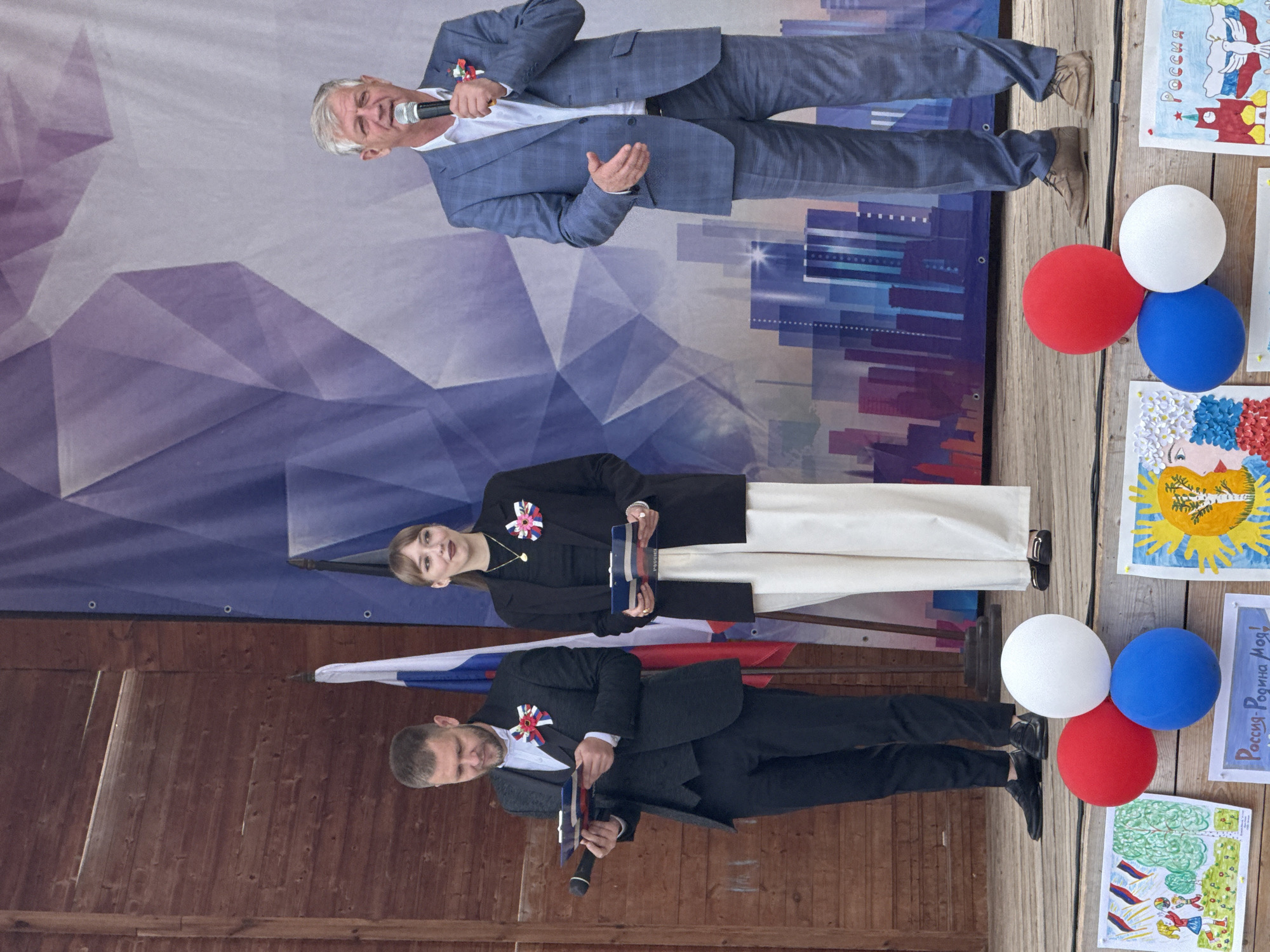 In Votkinsk, Russia, on a wooden stage, three individuals stand before a large banner depicting a geometric pattern in purple and blue against a light background, with a stylized city skyline at the bottom. On the left, a man in a dark suit and vest holds a clipboard. Next to him, a woman in a black blazer and white trousers also holds a clipboard. To the right, a man in a blue checkered suit speaks into a microphone. Russian flags are visible behind the two individuals on the left. In the foreground, three balloons – red, white, and blue – are arranged on the stage, alongside several colorful drawings, one of which appears to depict a portrait with flowers and the word "Россия" (Russia) written on it. Another drawing shows children holding flags and trees. The overall scene suggests an outdoor event or ceremony.