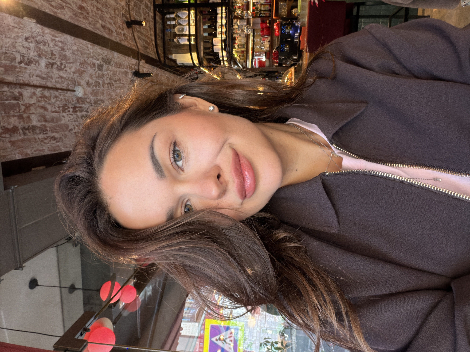 The image shows a medium-shot selfie of a woman with long, dark brown hair, taken indoors at a bar or restaurant. The woman is looking directly at the camera with a slight smile. She is wearing a brown jacket over a pink top and a silver necklace. She has light-colored eyes and pink lipstick. In the background, shelves are filled with bottles of alcohol, and a red bar counter is visible. The ceiling has exposed brickwork and industrial-style lighting fixtures, including some red spherical lamps. The environment suggests a modern and possibly trendy establishment. There is no specific information about the location in Saint Petersburg, Russia.