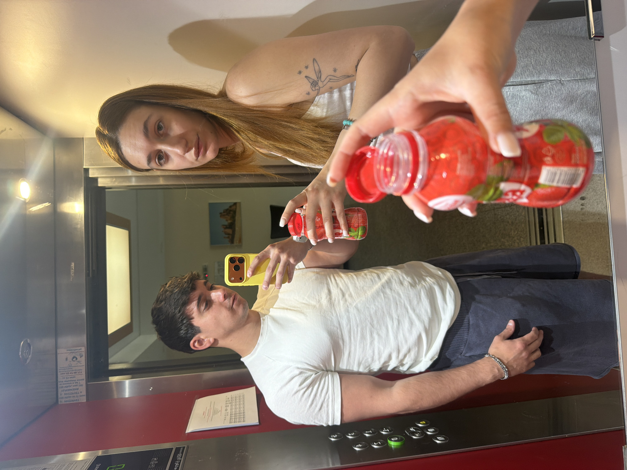 A mirrored selfie inside an elevator appears to be taken in Sofia, Bulgaria. A man in a white t-shirt and dark pants holds a yellow phone, presumably taking the photo. Reflected above him is a woman with long brown hair and a tattoo on her arm; her hand holds an open red strawberry-themed drink bottle. The elevator has red panels and buttons with Cyrillic characters, indicating the location. A document is visible on the elevator wall. A building interior is visible in the background.