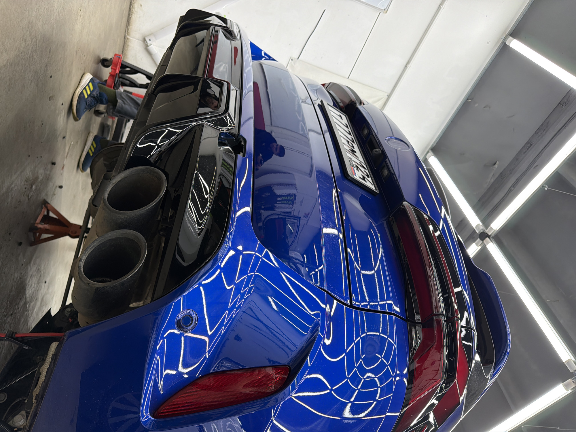 The image shows the rear of a blue car, possibly a BMW based on the license plate, in what appears to be a garage or workshop in Krasnoyarsk, Russia. The license plate is visible and bears the Russian flag. The car is elevated, possibly on a lift or jacks, as indicated by the presence of a red jack stand and a partially visible person with blue and yellow shoes underneath. The car has a custom black rear diffuser with quad exhaust pipes. The polished blue paint of the car reflects the overhead fluorescent lighting in a web-like pattern. The garage environment consists of concrete floors, a dark gray wall, and a white ceiling with mounted fluorescent tube lights.