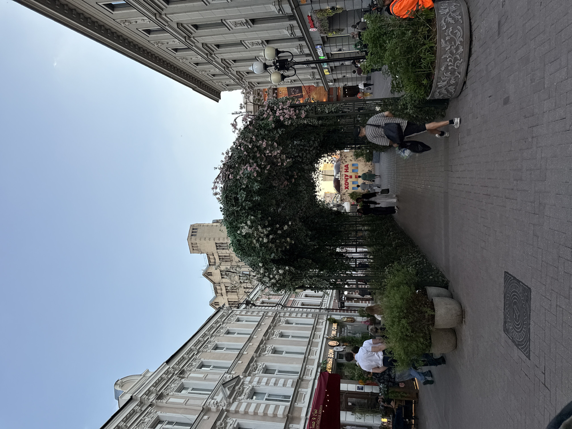 The image captures a vibrant urban street scene, likely a pedestrian zone, lined with multi-story buildings featuring classical architectural details. A large, ornate archway covered in dense greenery and pink flowers spans across the pathway, creating a focal point in the middle ground.
Several people are visible engaging in various activities. In the foreground on the right, a person wearing a striped shirt and shorts walks away from the viewer, carrying a black bag. Further down the street, near and within the archway, other individuals are strolling or gathered, some appearing to be seated at outdoor dining areas. A person in an orange vest is partially visible on the right side in the top portion of the image. The ground is paved with patterned grey interlocking bricks, and several planters containing bushes and small trees are placed along the street. Some planters are simple concrete, while others are intricately decorated metal. A circular manhole cover with a decorative pattern is visible on the right.
The sky is a clear, bright blue, suggesting good weather, and the soft lighting indicates either late afternoon or early evening. The architecture and visible text cues confirm the location context of Moscow, Russia.
Visible text includes:
* "АПТЕКА" (APTEKA), meaning Pharmacy, on a green sign above a shop.
* "ХОЧУ НА" (KHOCHU NA), meaning "I want to go to/on...", prominently displayed on a light-colored sign within the archway.
* "Уютный ДОМ" (Uyutny DOM), meaning "Cozy Home", and "BOWL CAFE" are visible on a red banner and signage for businesses on the left side of the street.