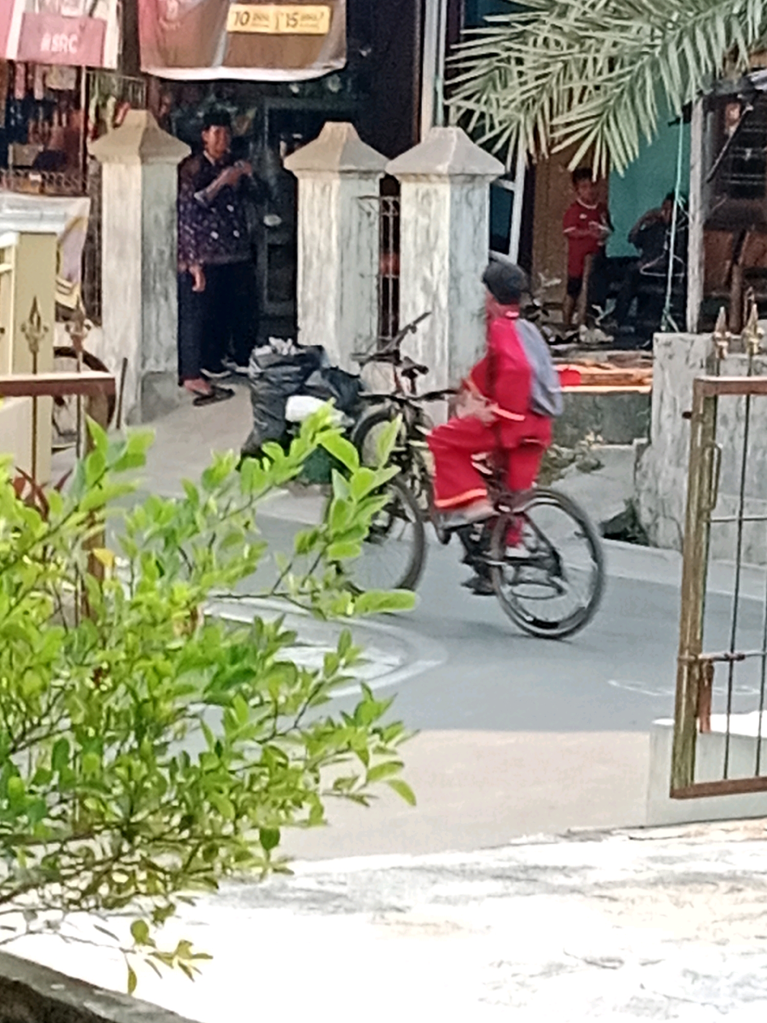 In Waru, Indonesia, a child wearing a red uniform rides a bicycle, facing away from the camera. The child is also wearing a dark cap and a gray backpack. The bicycle is dark in color. In the background, a person in traditional clothing stands under an awning, looking towards the right. Behind them, another child in a red shirt and shorts stands near a doorway. A palm tree is visible above the scene, and green foliage is in the foreground.