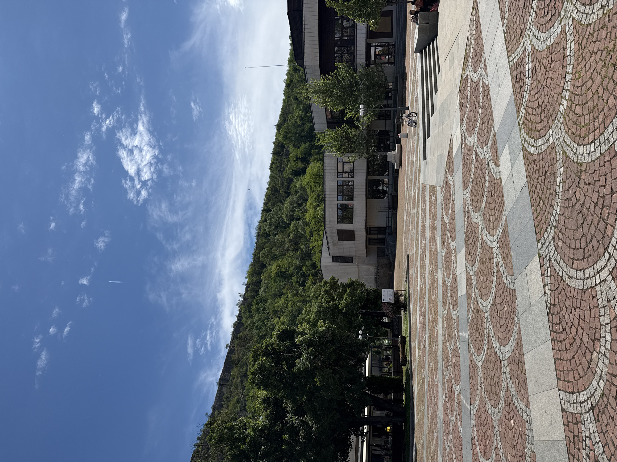 The image captures an outdoor scene in Lovech, Bulgaria, featuring a patterned stone square and the urban environment around it. The foreground shows a pavement with distinctive semi-circular patterns composed of reddish-brown and gray stones.
The background displays a tree-covered hillside that appears quite steep, typical of the geography around Lovech. A building with a modern design is situated near the base of the hill. Above the hill, the sky is blue with scattered clouds. A contrail left by an airplane is visible across the sky.
A row of trees is planted along the edge of the pavement, creating a natural separation between the square and the hill. People are present on the stairs leading up to a building. Street lights and benches are present along the edge of the sidewalk.