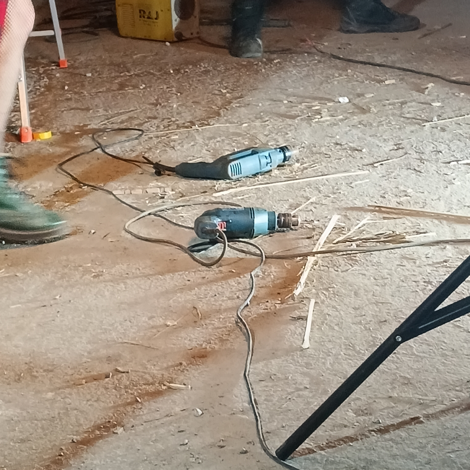 The scene appears to be the interior of a workshop or construction site, judging by the wood shavings and tools scattered on the floor. Two electric drills are lying on the concrete floor, each with a power cord. In the background, there is what looks like a yellow portable generator and a stepladder. The image shows at least two people, partially visible by their feet and lower legs. A black tripod leg is visible in the bottom right corner of the scene. The location is unknown and cannot be deduced from the image. Therefore, providing a city location such as Panaji, India is not possible.