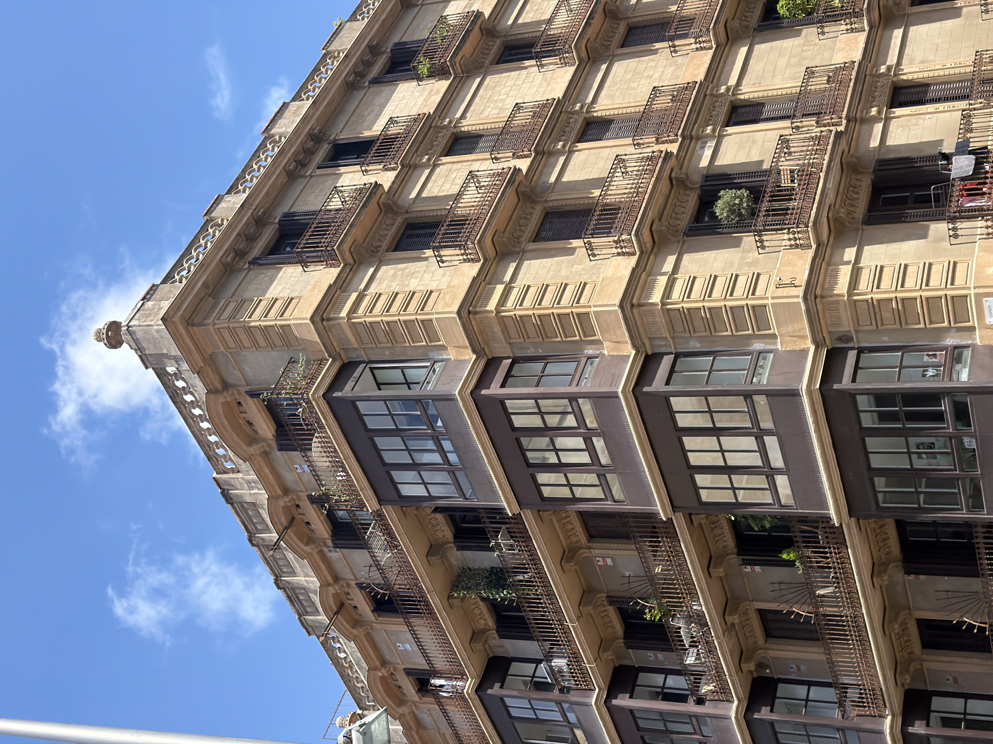 This is a photograph of the facade of a building with a light brown or beige exterior. The building has multiple stories, each with numerous windows and balconies. The balconies are ornate with wrought-iron railings and some have plants. The windows are also dark-framed and appear to be large, with some having shutters. The building has decorative architectural details, including cornices and moldings. The photograph is taken from a low angle, looking up at the building, and the sky, which is blue with some white clouds, is visible above. The sunlight suggests it is daytime. The image is rotated, making the building appear to tilt.
There are no discernible people in the image. Objects visible include plants on the balconies, some laundry hanging on a balcony, and architectural features of the building. The scene is an urban setting, likely a city street in Barcelona, Spain, given the architectural style. There is no specific activity or event taking place, it's a static view of the building. Notable details include the repetitive pattern of windows and balconies, the decorative stonework, and the contrast between the building's facade and the blue sky. The time of day appears to be sunny and bright. There is no visible text in the image.