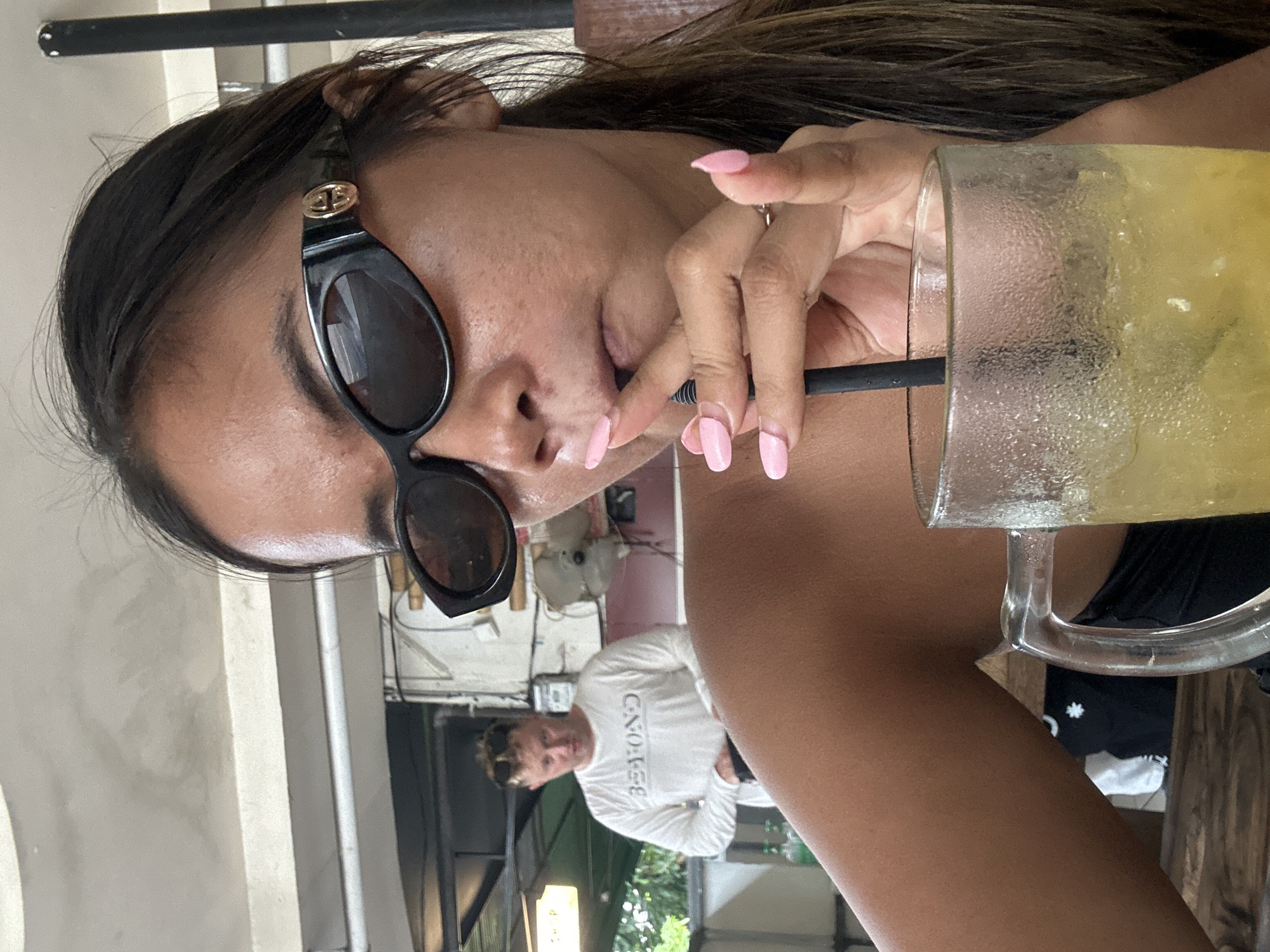 In Kuta, Indonesia, a woman with long brown hair and sunglasses is taking a selfie while holding a glass of yellow drink with ice and a straw. Her fingernails are painted pink. In the background, a man wearing a white long-sleeved shirt with the word "BEYOND" printed on it is visible. The environment appears to be an outdoor seating area with a ceiling fan and a white wall.
