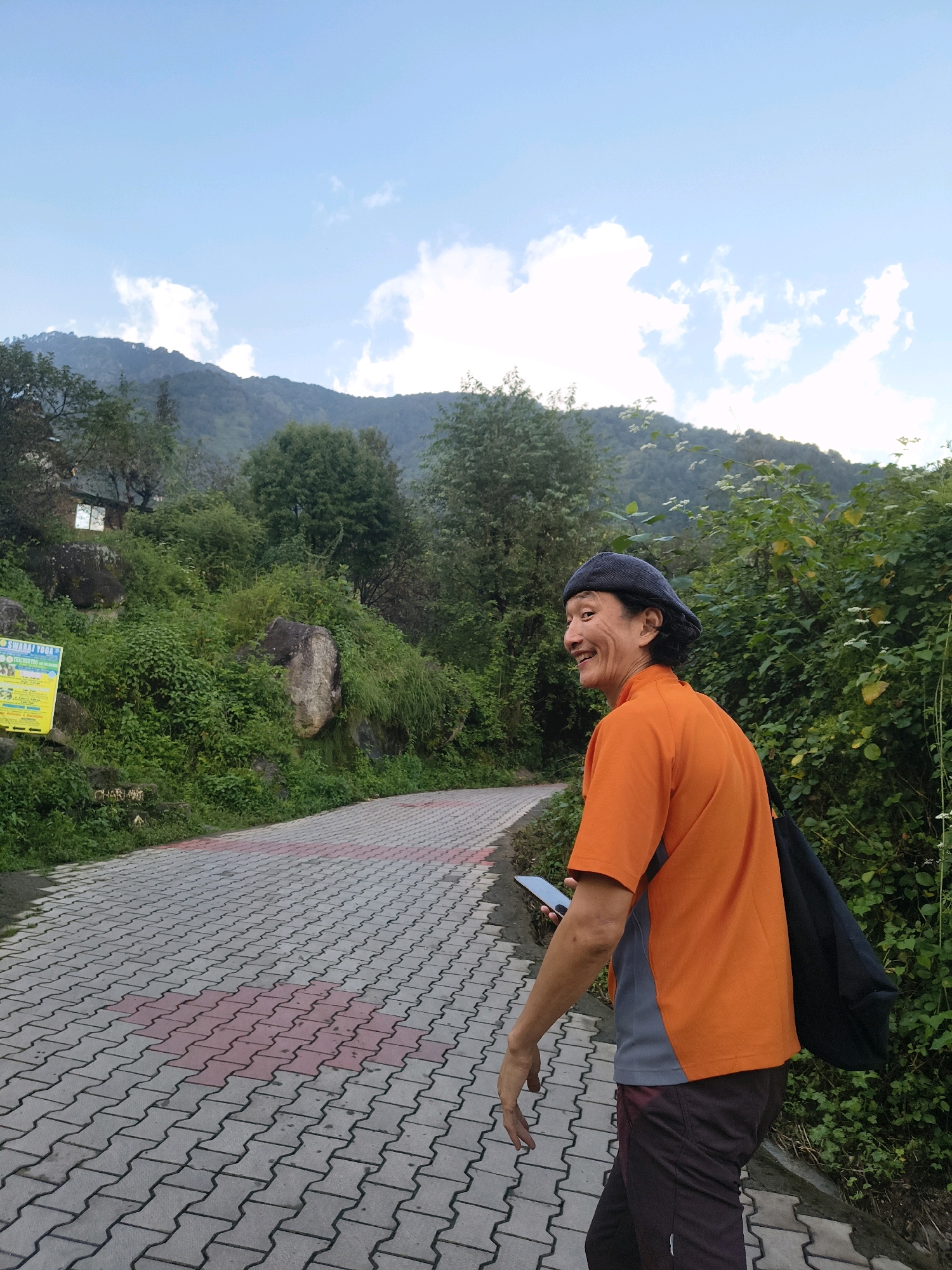 A man wearing a blue beret, an orange shirt, and dark pants walks on a paved path. He is smiling and looking over his right shoulder. He holds a phone in his left hand. The path is made of interlocking pavers with red accents. The man is walking in front of a lush green hillside with trees and rocks. In the background, mountains are visible under a partly cloudy sky. A yellow sign is posted on the left side of the path. The scene appears to be outdoors, likely in a natural or semi-rural setting. The man's attire and the overall environment suggest a casual outdoor activity. The presence of the paved path indicates a developed area, possibly a park or a walking trail. The man's relaxed posture and smile suggest he is enjoying himself. The question does not provide the location in Haini, India.