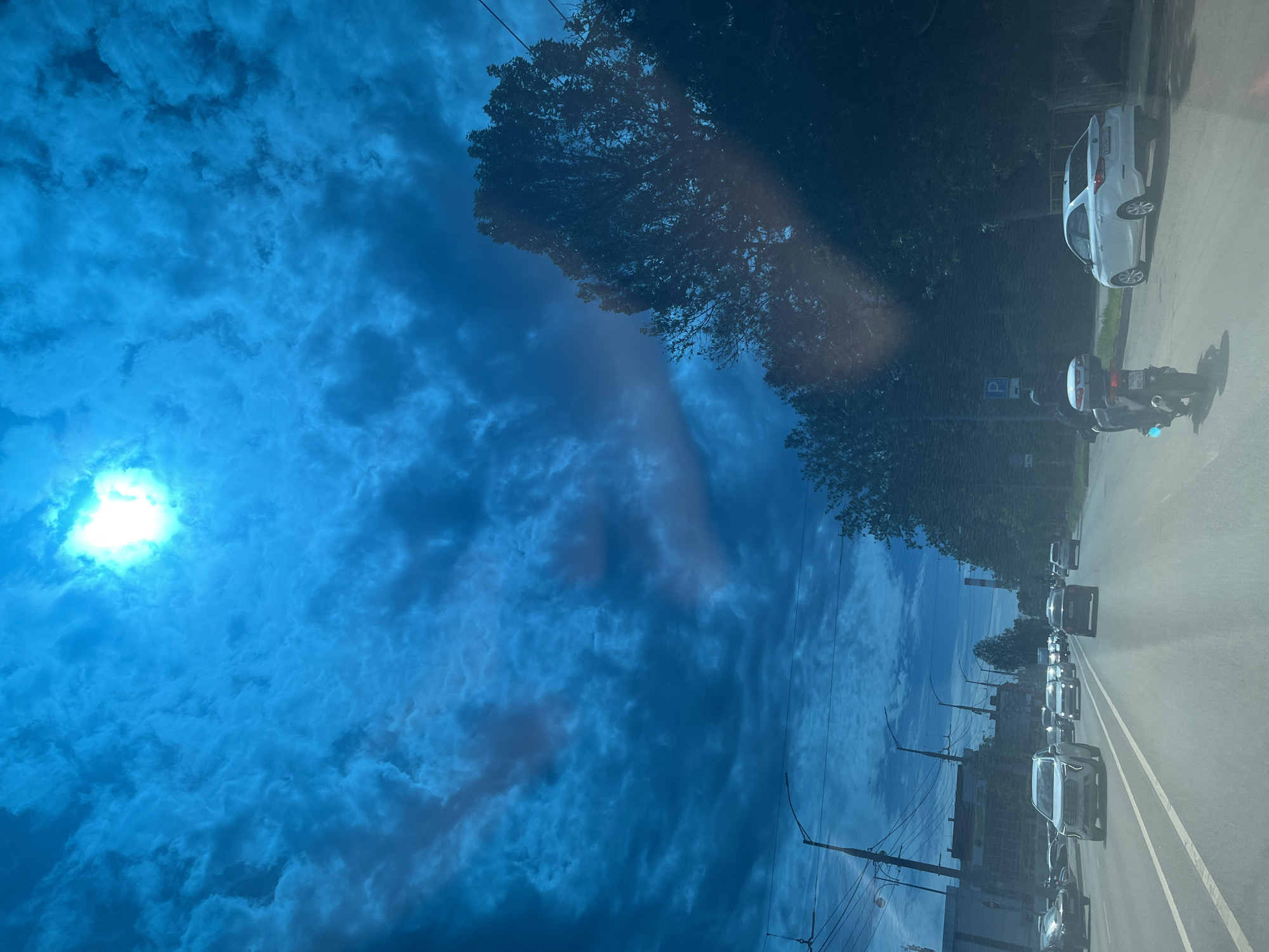 The image, taken from inside a vehicle, captures a daytime street scene under a vivid blue sky. The photo is rotated 90 degrees clockwise; when viewed upright, the sky dominates the upper and left portions, while the road and vehicles are on the right and bottom.
A bright, almost blinding light source, likely the sun or its reflection, is visible in the upper left, casting a strong glare. The sky is filled with numerous clouds, ranging from wispy white to denser grey formations, providing a dynamic backdrop.
Along the right side of the image (top if rotated correctly), a dense line of dark green trees borders an asphalt road. Utility poles and power lines are visible, indicating an urban or suburban setting. The road itself appears to be multi-laned, with visible white lane markings.
Several vehicles are present on the road. In the foreground, a white car, possibly a hatchback or small SUV, is visible, alongside a motorcycle with a rider. The motorcycle's license plate is partially legible, starting with "A488". A blue parking sign with a white 'P' symbol is visible behind the motorcycle. Further down the road, a line of cars indicates traffic. Reflections on the window through which the photo was taken are evident, particularly on the road surface and some vehicles, layering elements of the sky onto the scene.
The image depicts a typical street environment in what is identified as Krasnodar, Russia, during a partly cloudy, sunny day.