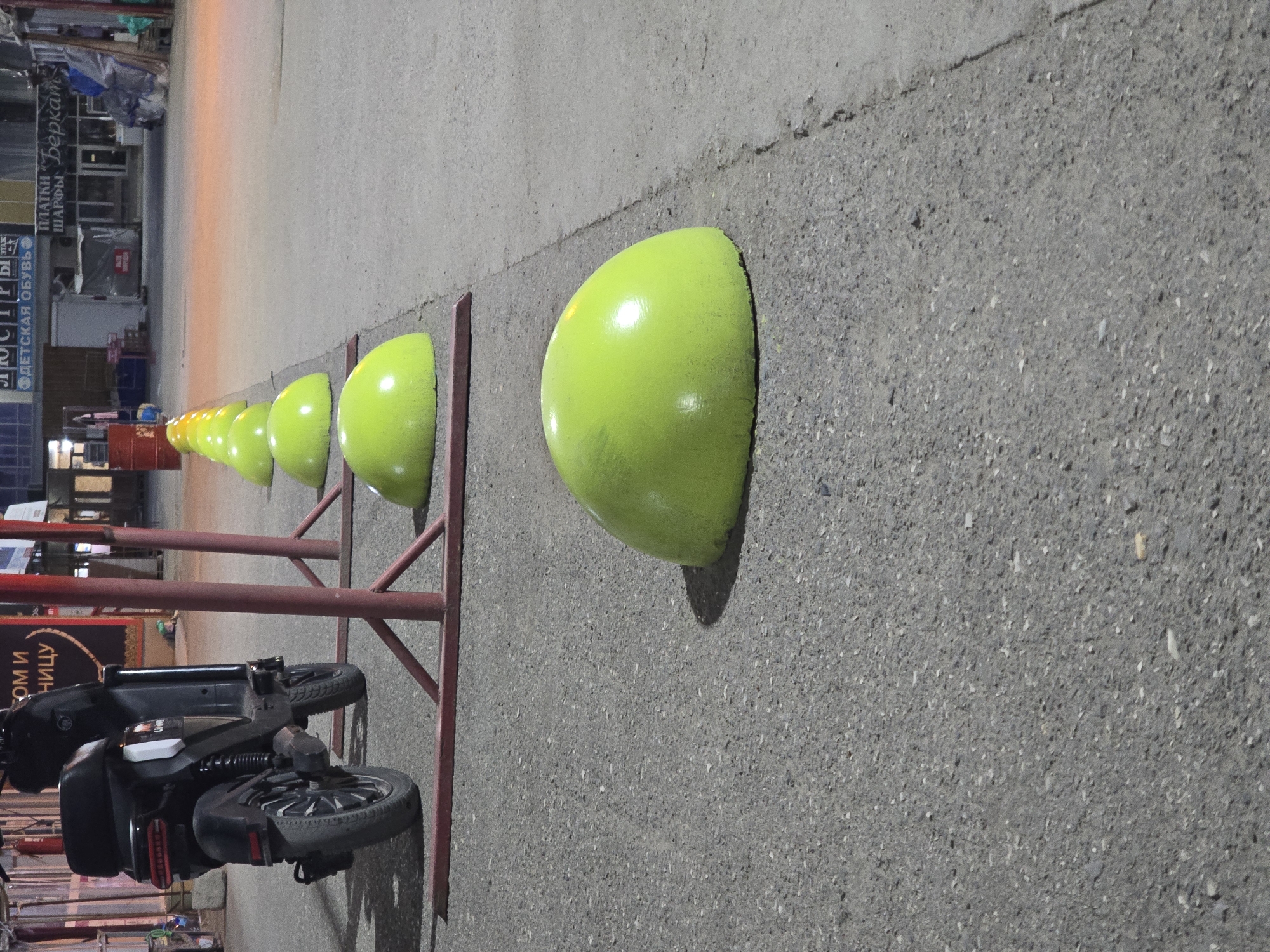 This media file shows a line of bright green, rounded bollards set into asphalt. The bollards are spaced evenly and appear to be a form of traffic calming or pedestrian barrier. In the background, a red metal fence runs parallel to the bollards, with a dark-colored electric scooter parked beside it. Further in the background, there are glimpses of buildings and storefronts, suggesting an urban or commercial setting. The lighting suggests it might be evening or a dimly lit area, with artificial light sources visible reflecting off the pavement.
Some visible text in the background is in Cyrillic script. On one storefront, the words "ДЕТСКАЯ ОБУВЬ" (Children's Shoes) and "ПЛАТКИ ШАРФЫ" (Scarves) are partially legible. Another sign reads "СОМ И" (Some I) and "НИЦУ" (Nitzu).
The scene appears to be set in Khasavyurt, Russia. The overall impression is of a functional outdoor space, possibly a sidewalk or pedestrian zone, with practical elements like bollards and a fence for crowd control or safety. The presence of the scooter and storefronts indicates a connection to daily life and commerce.