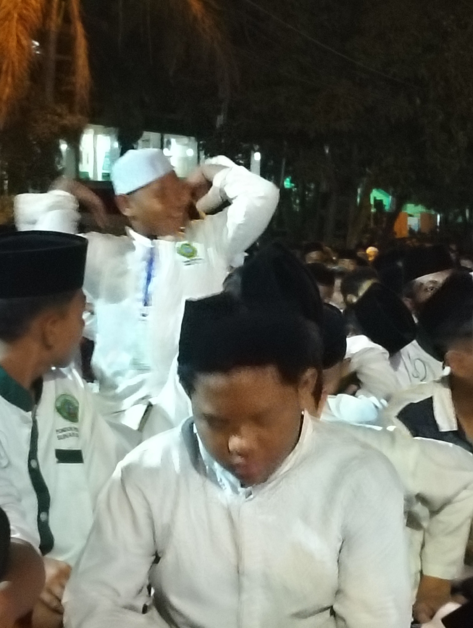The image depicts a crowd of people, predominantly young males, gathered outdoors at night. Many individuals are wearing white Islamic attire, including peci (caps). In the foreground, a young person with dark hair is wearing a white shirt and looking down. To the left, another individual in a black peci and a white and green collared shirt is visible, facing towards the center of the crowd. In the background, some individuals are wearing white outfits and peci, with one person adjusting their cap. Lights are visible in the background, suggesting an event or gathering in a public space. The environment appears to be a dimly lit outdoor area, possibly a field or courtyard, with trees and buildings faintly visible. The context provided places the scene in Drajat, Indonesia, suggesting a local event or religious gathering.