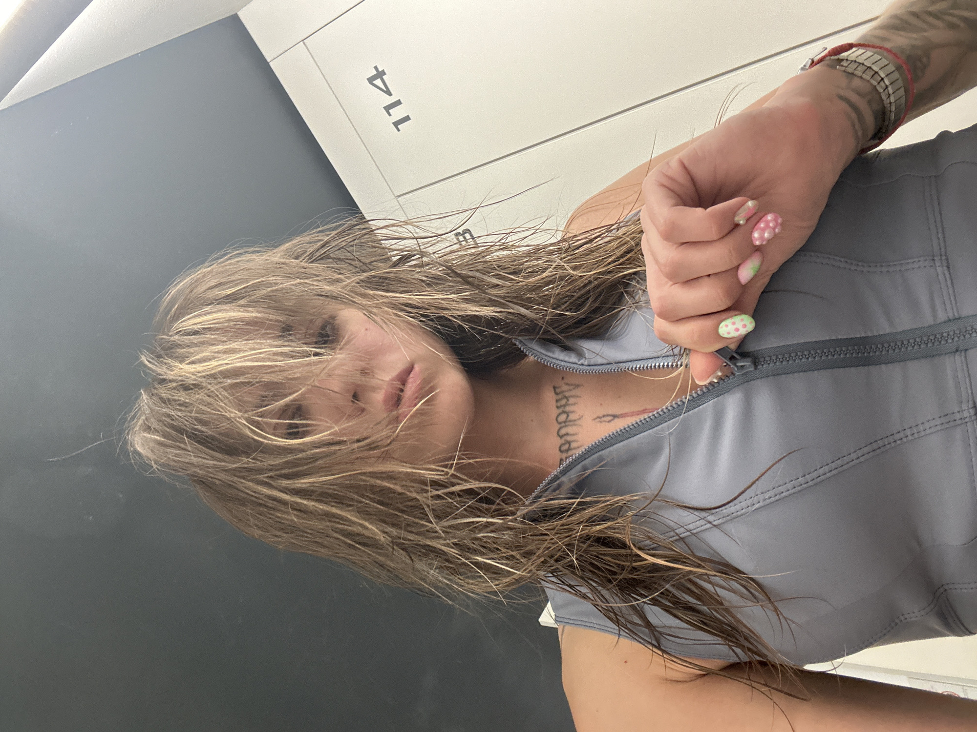 A low-angle medium shot captures a woman with wet, blonde hair in what appears to be a locker room, possibly in Moscow, Russia. She is wearing a grey, sleeveless, zippered top. Her hair is falling over her face, partially obscuring her features, but her lips are visible and slightly parted. A tattoo is visible on her chest. Her right hand is raised to the zipper of her top, and her fingernails are painted with a pink and green speckled design and some have raised pink dots. A silver watch with a metal band is on her left wrist, along with a thin red string bracelet. To the right of the woman, a white locker door is visible, with the number "114" printed in black at the top. The background behind her is a dark grey wall.
