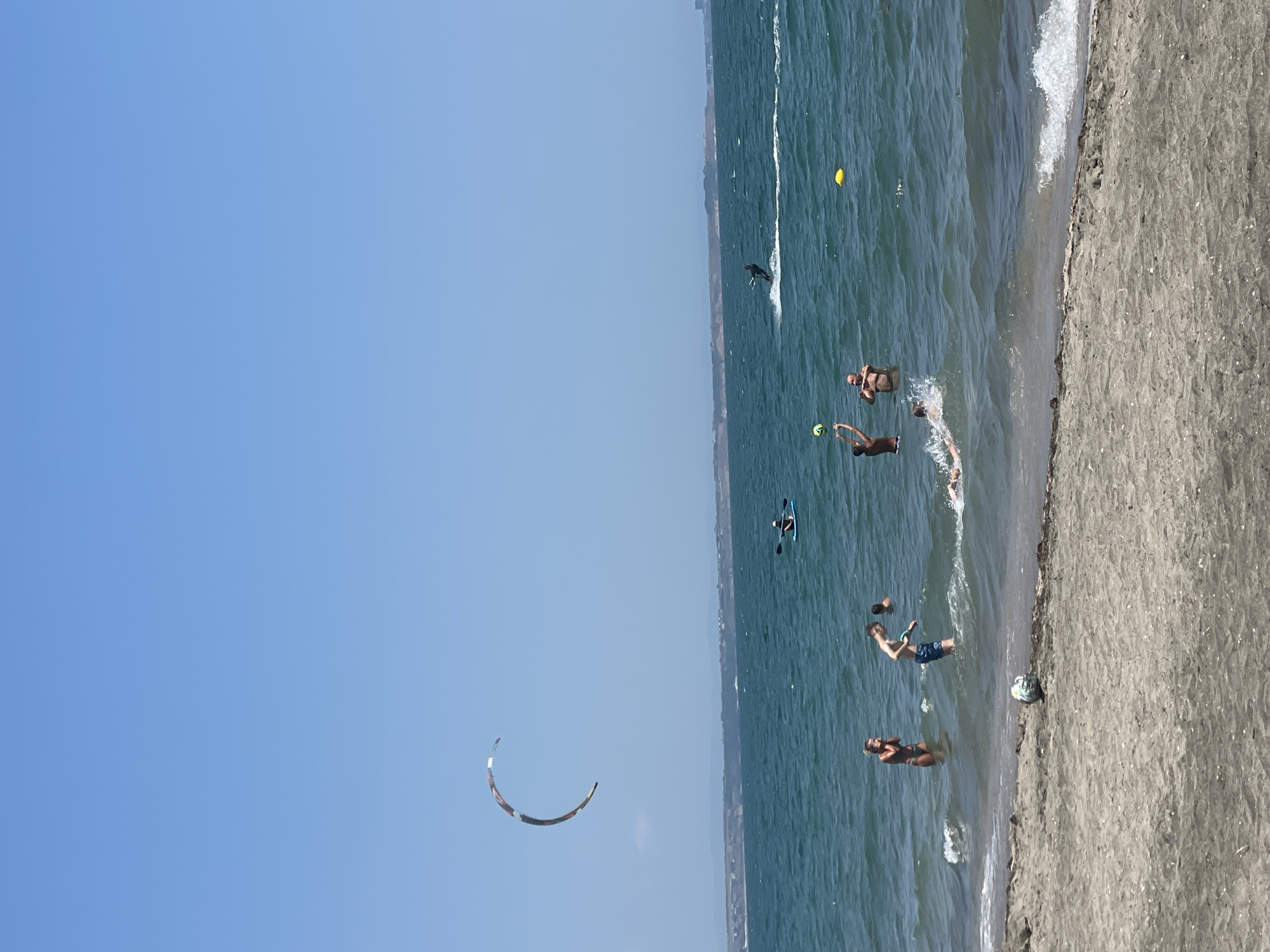 The image depicts a daytime scene at a beach, likely located in Burgas, Bulgaria, given the context. The sky is a clear, bright blue. The water is a deep teal color, with moderate waves breaking near the shore. The shoreline is composed of sand, with some scattered debris.
In the water, several individuals are engaged in various activities. In the foreground, a few people are standing in shallow water. To the left, a woman in a dark bikini is looking towards the water. Near the center, a young boy in blue swimming trunks is holding a green object. Further out, a person is lying on their back in the water. In the middle ground, two men are standing in the water, with one appearing to be playing with a ball, throwing it upwards. A yellow buoy floats in the water to their right.
In the background, on the water, there are several watercraft and activities. A person is seated on a kayak, paddling. Further to the right, a kiteboarder is actively riding the waves, with a spray of water behind them. High in the sky, a large kite, possibly for kitesurfing, is visible, curved against the blue backdrop. A distant shoreline with hills and some buildings is visible on the horizon.