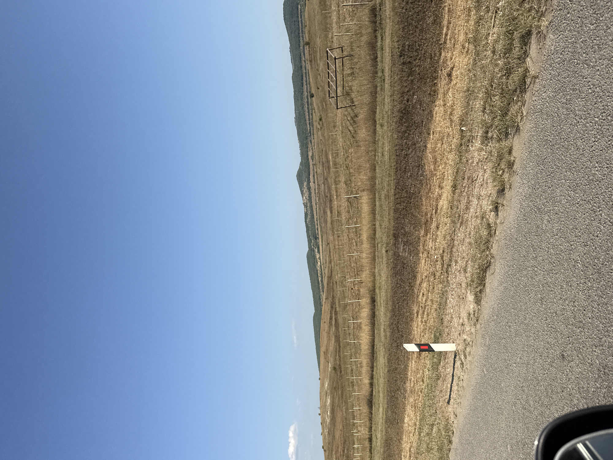 The image depicts a rural roadside scene on a clear, sunny day. A paved road runs diagonally across the lower left corner of the frame, with the side mirror of a vehicle visible in the extreme lower left. A white and red reflective road marker is positioned on the right side of the road. Beyond the road, a dry, grassy field stretches towards rolling hills in the distance. The field is sparsely dotted with thin, white posts, possibly part of a fence line, and a simple wooden structure, resembling a hay rack or shelter, is situated in the middle ground. The sky is a clear, bright blue with a small, white cloud near the left horizon. The landscape appears to be agricultural or undeveloped land, with a sense of open space and a dry, summery or autumnal atmosphere. The location is identified as Почтовское сельское поселение, Ukraine.
