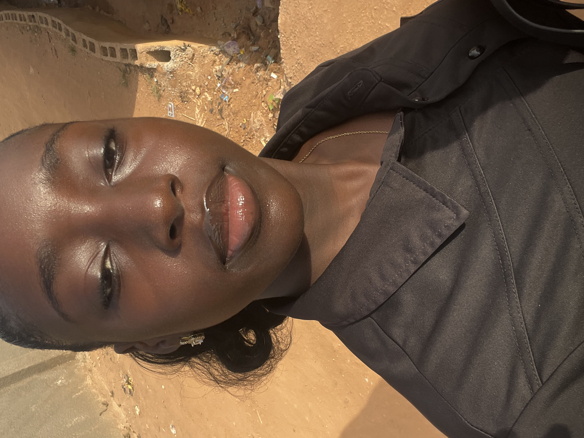 A portrait-style image, taken outdoors in Abuja, Nigeria, features an adult individual, likely female, with dark skin, positioned to face the camera. The person is wearing a black collared top or jacket and a thin gold chain necklace. A sparkling earring with a rectangular-cut stone is visible on their right ear. Their lips have a glossy appearance, and their skin appears radiant with natural sheen under direct sunlight.
The background consists of an unpaved, dusty brown ground with visible debris and litter, including small pieces of plastic and paper. A low wall made of concrete blocks is partially discernible behind the person. The bright lighting suggests it is daytime with clear, sunny weather, casting subtle shadows and highlighting the subject's features and clothing. The individual's expression is neutral, appearing calm. There is no visible text in the image.