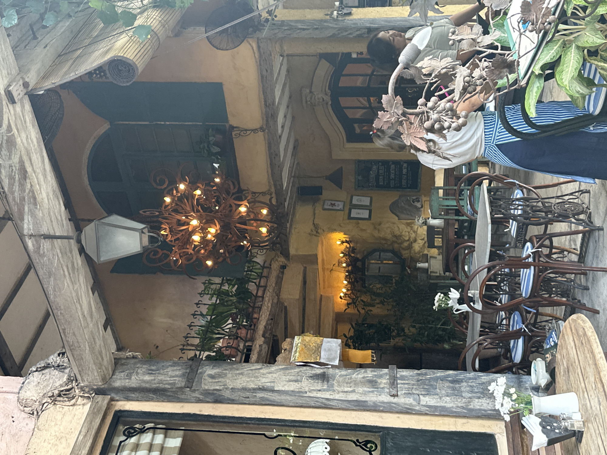 The image depicts the outdoor seating area of a restaurant or cafe, likely located in Ubud District, Indonesia. The architecture features rustic elements with exposed wooden beams, textured walls in earthy tones, and decorative wrought iron accents. A large, ornate chandelier hangs from the ceiling, illuminating the space with warm light.
Several tables with chairs are arranged for dining. Some chairs appear to be antique sewing machine bases, adding a unique vintage touch. The tables are set with white napkins and salt and pepper shakers. One table features a vase of white flowers.
Two women are visible in the image. One woman, wearing a white top and a blue and white striped apron, is standing near a table, possibly a server. She is partially obscured by a decorative metal plant structure. Another woman, wearing a light-colored top, is also near a table, and her body is mostly out of frame.
In the background, there are glimpses of more seating, potted plants on a railing, and arched doorways. The overall atmosphere suggests a charming and eclectic dining establishment.
