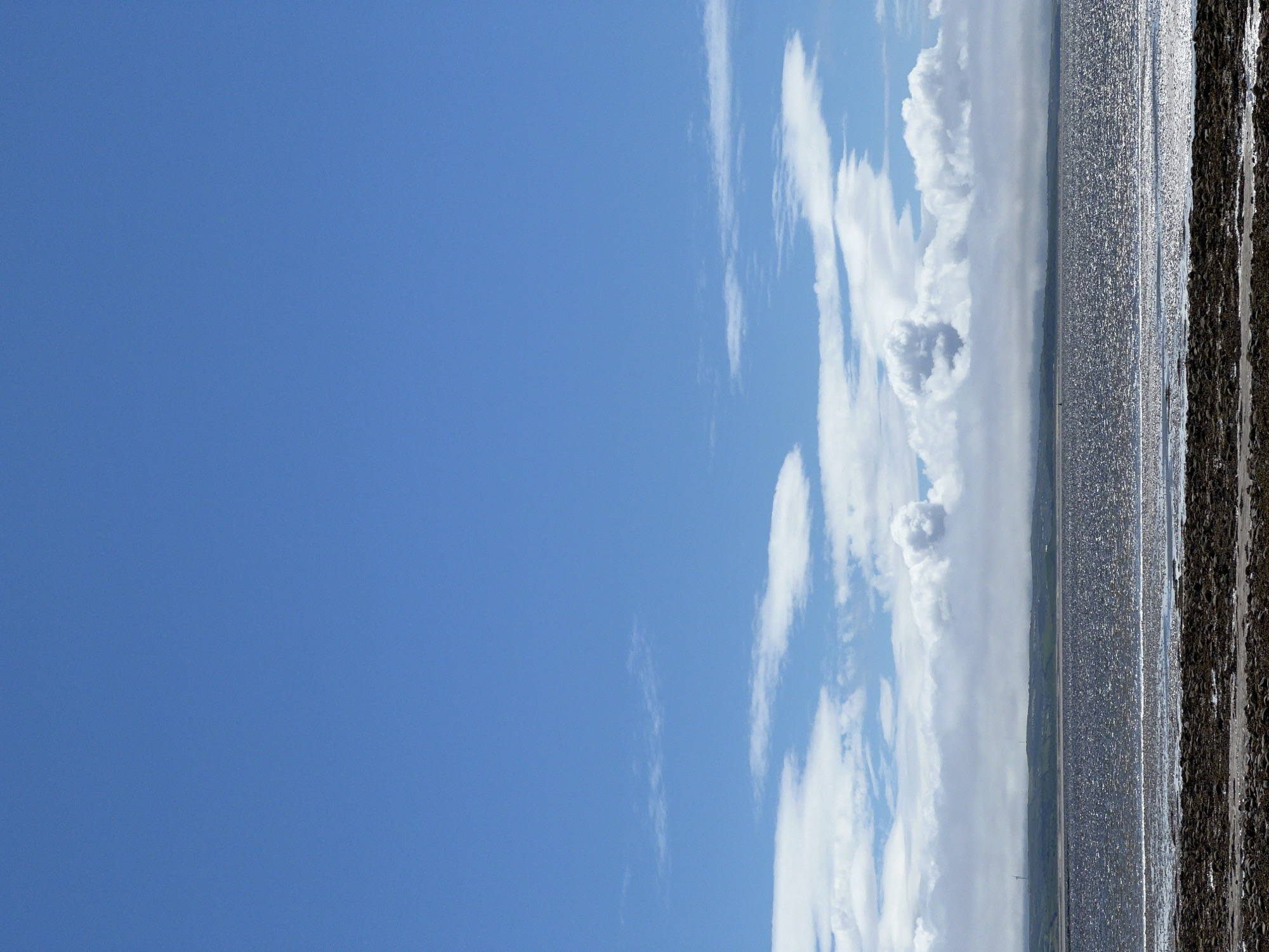 An outdoor, daytime, vertical-format, eye-level shot shows a broad expanse of water with a shoreline in the foreground and a distant landmass under a bright blue sky with scattered clouds. The shoreline is composed of dark, wet rocks and pebbles. Beyond the rocks is a body of water that appears to be reflecting the sky and sunlight, creating a shimmering effect. A low-lying, green landmass with gentle hills is visible on the horizon, partially obscured by a band of atmospheric haze or fog. The sky is a vivid blue, with large, fluffy white clouds dominating the upper portion of the image, and thinner, wispy clouds lower down, closer to the horizon. There are no people or animals visible in the image. The scene appears to be a coastal landscape. The location is the city of Mountcharles, Ireland.