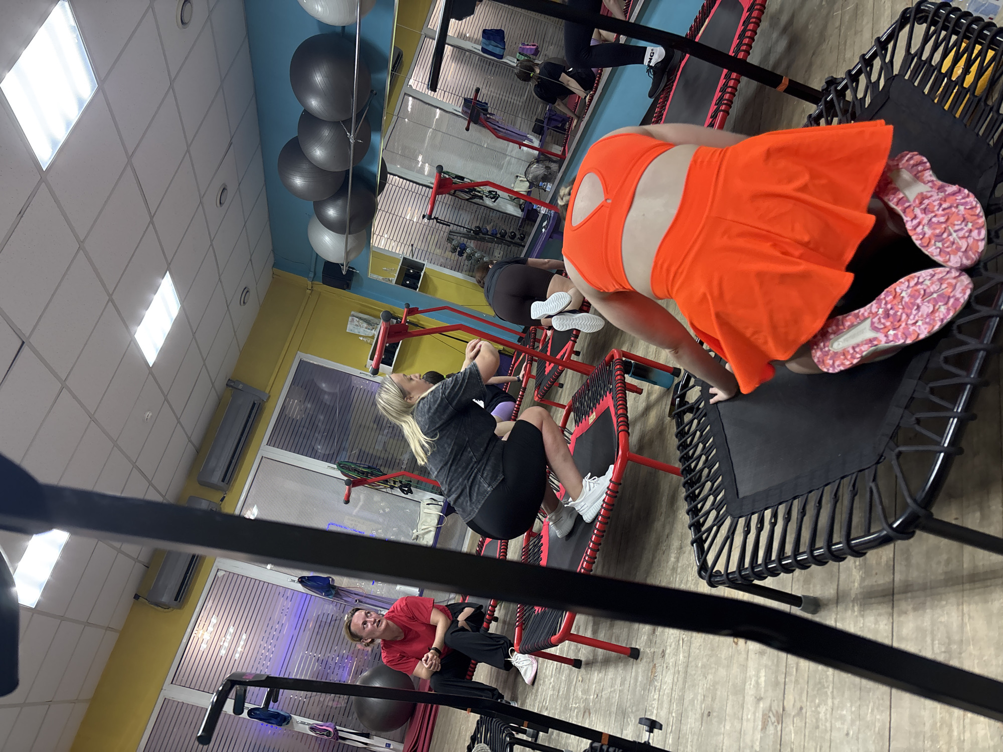 A group of women are engaged in a fitness class at a gymnasium. The women are using individual mini-trampolines. The room is brightly lit with fluorescent lighting and has large mirrors on one wall. There are exercise balls on a shelf. The floor is made of wood. The overall impression is one of an active and modern fitness studio.