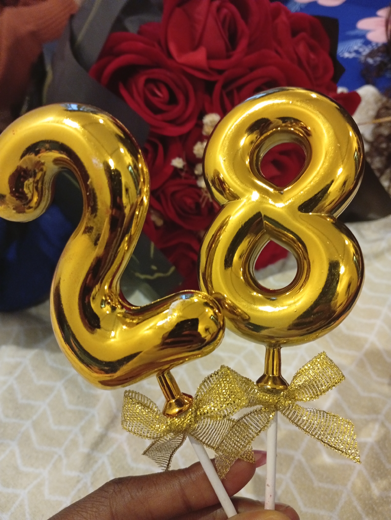 This media file features a close-up of a hand holding two golden, inflatable number balloons that spell out "28". The balloons are reflective and appear to be made of a shiny material, possibly Mylar. Each balloon is attached to a white stick and decorated with a small, golden, glittery bow.
In the background, out of focus, are several red roses, possibly part of a bouquet or floral arrangement. The overall color palette is dominated by gold, red, and muted tones of the background fabric. The lighting suggests an indoor setting, likely artificial light given the bright reflections on the balloons. The hand holding the balloons is dark-skinned, with painted fingernails.
The scene is likely related to a birthday celebration or anniversary, given the prominence of the number "28". The presence of the balloons and flowers suggests a festive occasion. No specific time of day or weather conditions are discernible from the image. The location context provided is Abuja, Nigeria, but there are no explicit visual cues within the image to confirm this. There is no visible text in the media file.