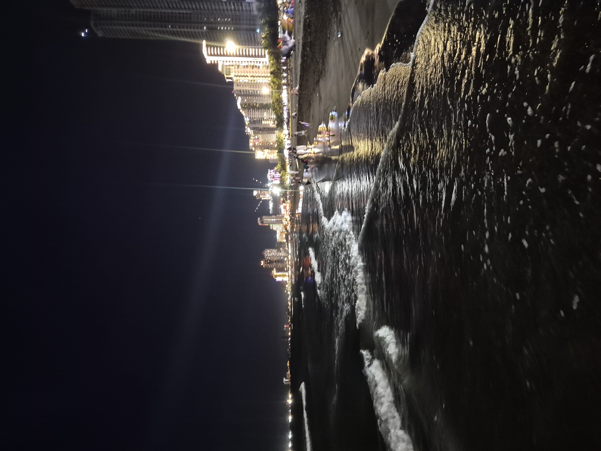 This is a night time, low-angle, long exposure photograph of the beach in Da Nang, Vietnam, with the city skyline in the background. The foreground is dominated by the dark ocean with white waves lapping at the shore. The sand is dark and wet from the tide. In the mid-ground, people are walking along the beach, their reflections shimmering on the wet sand. The background features a brightly lit city skyline with tall buildings illuminated by numerous lights. The sky is dark, suggesting it is nighttime. The overall impression is one of a vibrant, active beach scene at night.