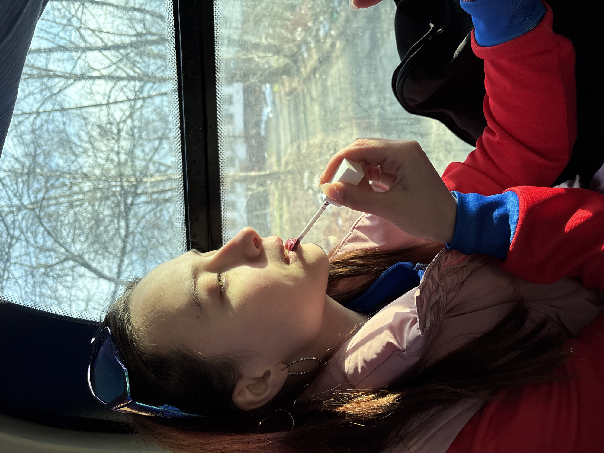 A young woman is shown applying lip gloss in front of a window. The woman is wearing a red and blue jacket over a pink puffy jacket. She is wearing silver hoop earrings. Her brown hair is partially illuminated by sunlight. Outside the window, there are bare tree branches and buildings. The city is unidentified.