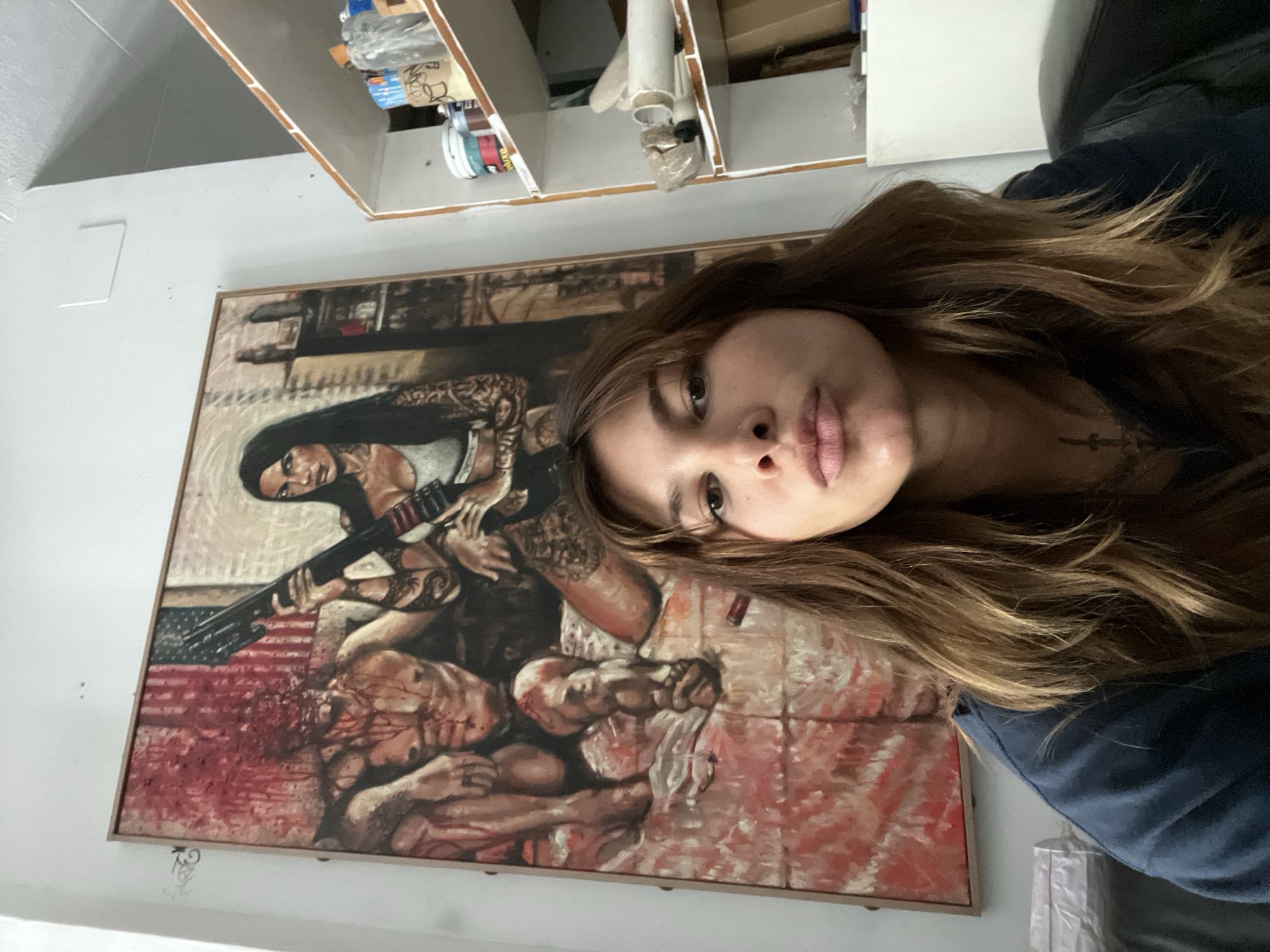 A medium shot captures a young woman with long, wavy brown hair and light skin in the foreground, looking directly at the camera with her lips slightly pursed. She is wearing a dark blue long-sleeved shirt, and a tattoo is visible on her chest. Behind her, a large, framed painting dominates the background. The painting depicts two figures against a backdrop that includes an American flag. On the left, a muscular male figure is seated and appears to have severe head trauma with blood splattered around him. On the right, a woman with dark hair, tattoos, and a white top is holding a rifle. To the right of the painting, a white shelf unit is visible, filled with various items, including paint cans, rolls of paper, and other supplies. The environment appears to be an indoor space, possibly an artist's studio or a home, with a plain white wall. The overall scene is a selfie taken in front of a striking and graphic piece of artwork. The location is not specified in the image or its context.